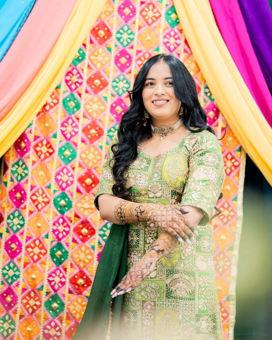 A woman in a green traditional dress with intricate patterns, wearing jewelry and henna on her hands, stands in front of colorful fabric drapes and a vibrant decorative backdrop. Mehndi bridal shoot