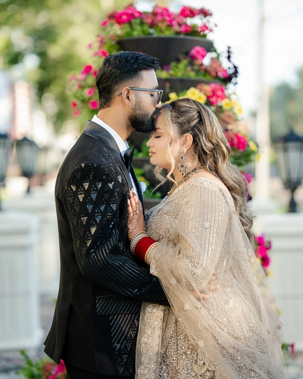 A couple dressed in wedding attire standing close together outdoors, with the man kissing the woman's forehead, surrounded by colorful flowers.