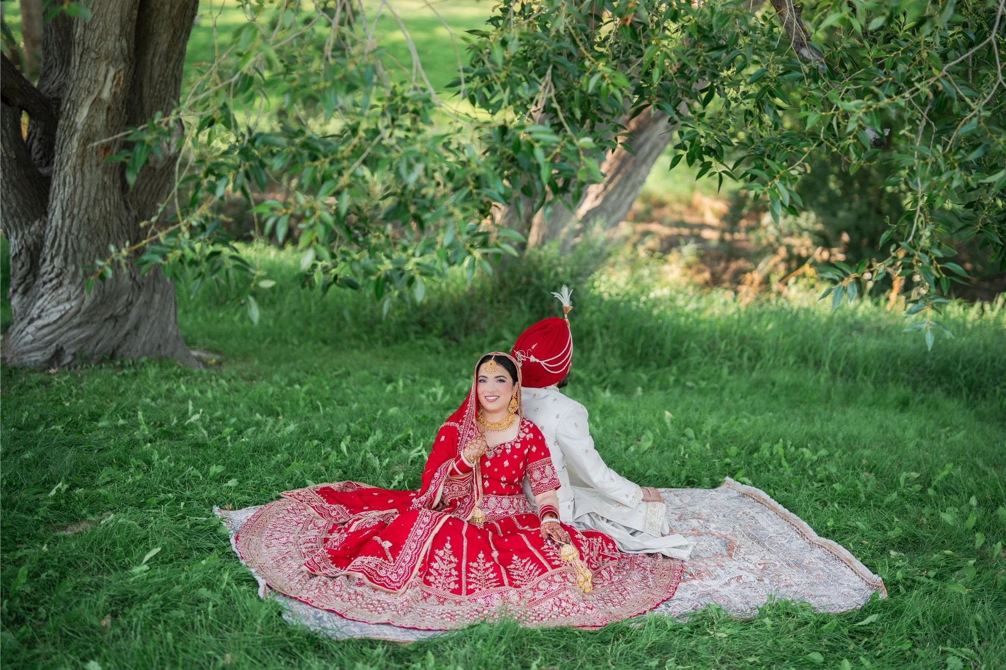 A bride dressed in a red traditional wedding outfit with gold jewelry sits on a patterned rug on the grass, smiling, with her back to a groom in a white outfit and red turban, under a large tree in a lush green outdoor setting.