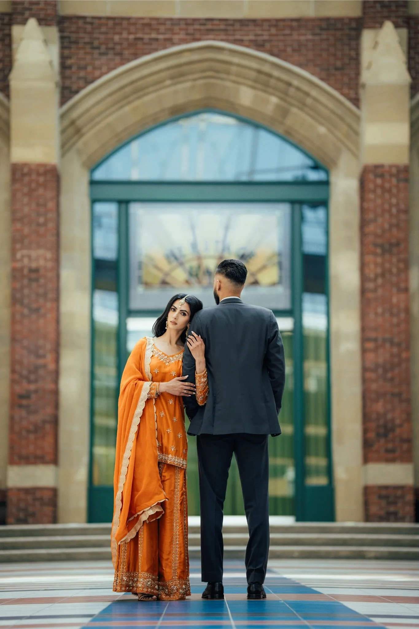 A woman and a man in formal attire standing on steps in front of a large arched glass door, with the woman wearing an orange traditional outfit and the man in a black suit.