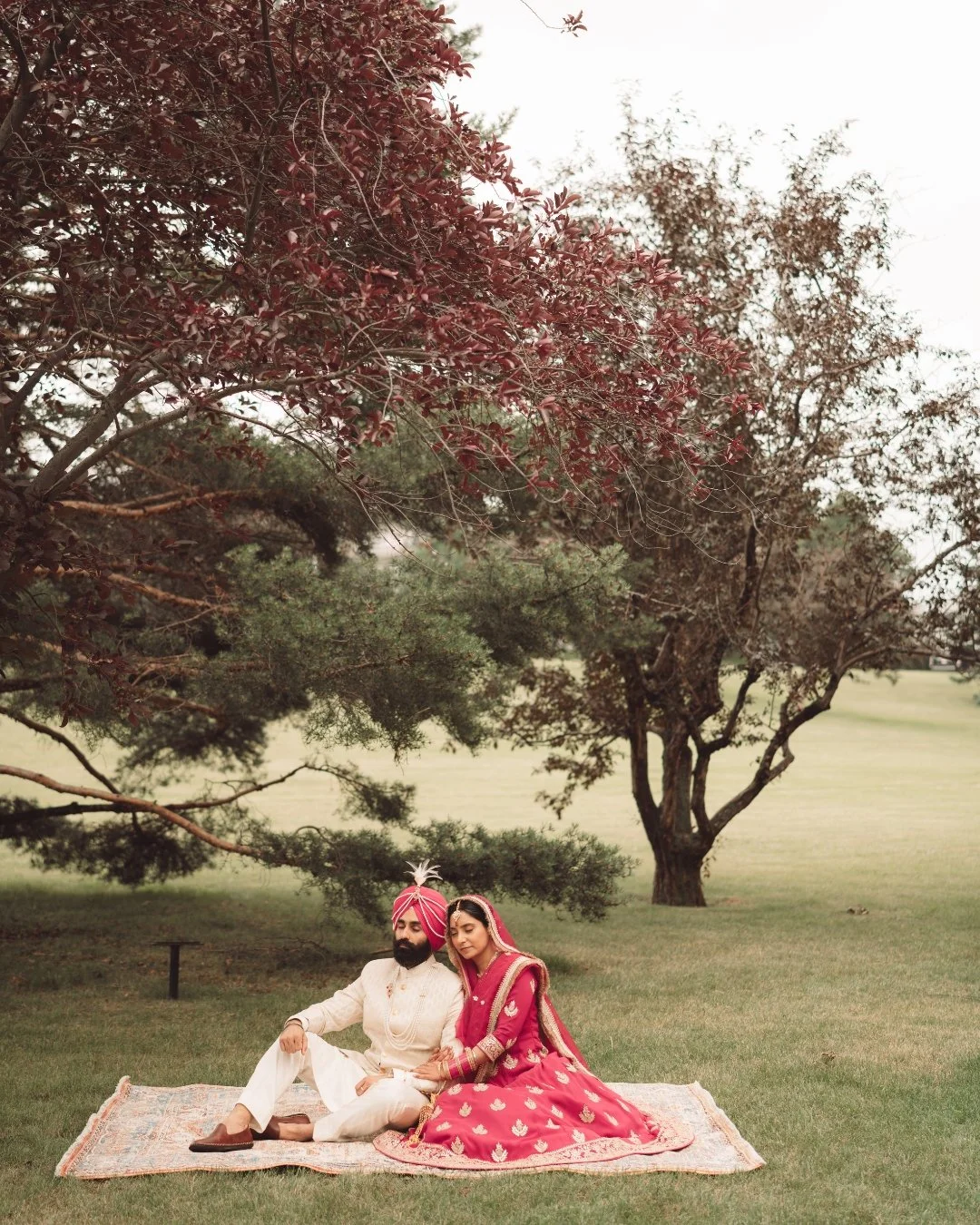 A couple dressed in traditional Indian wedding attire sitting on a rug outdoors under a large tree.