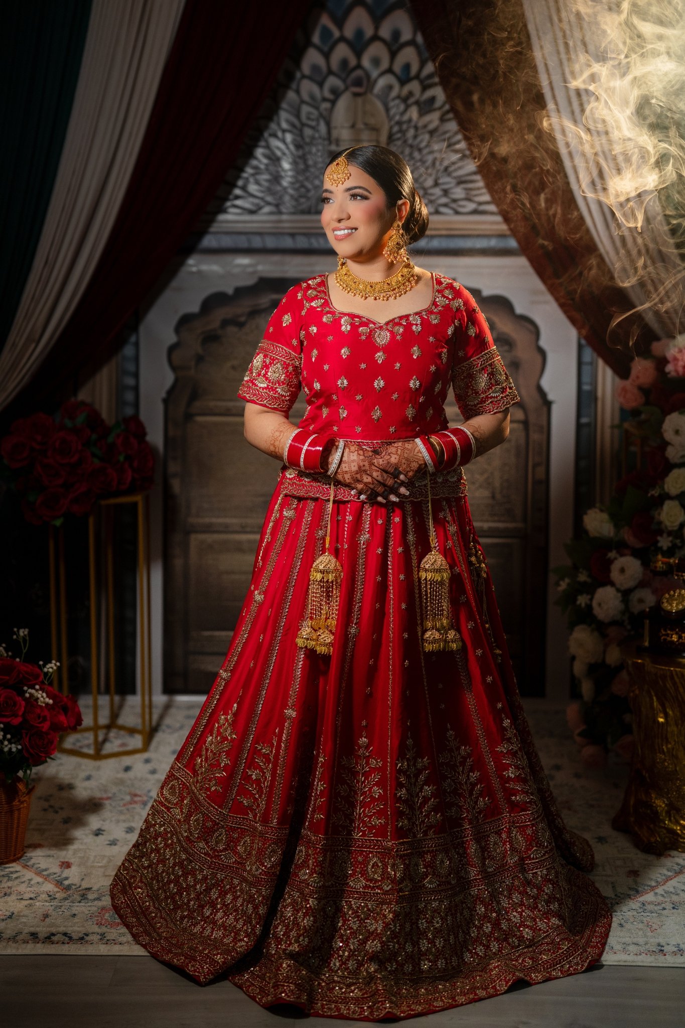 A woman dressed in a traditional red Indian bridal outfit, standing by a decorated wedding venue, with henna on her hands, gold jewelry, and intricate embroidery on her dress.