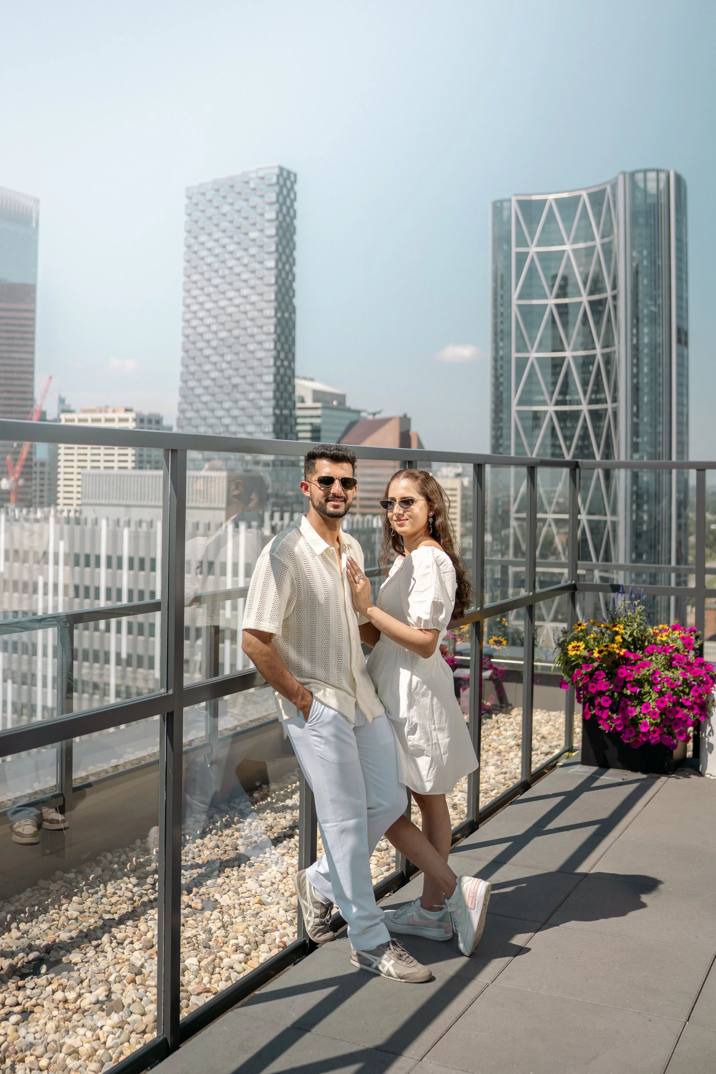 A man and woman stand on a rooftop terrace with city skyline in the background. They are wearing casual white clothes and sunglasses, posing together near a glass railing with flower pots.