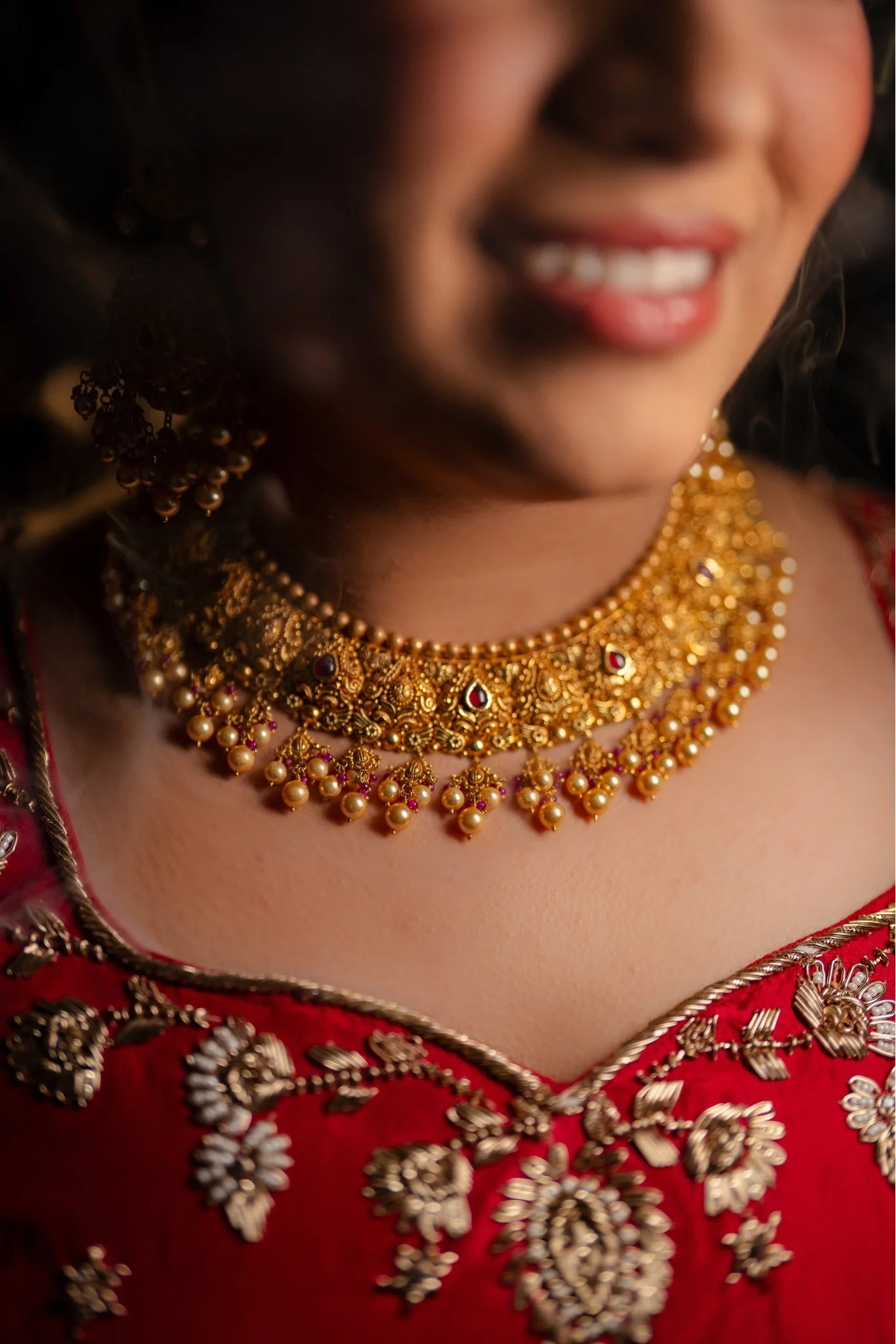 Close-up of a woman wearing traditional red and gold embroidered clothing, gold jewelry, and showing a smile.