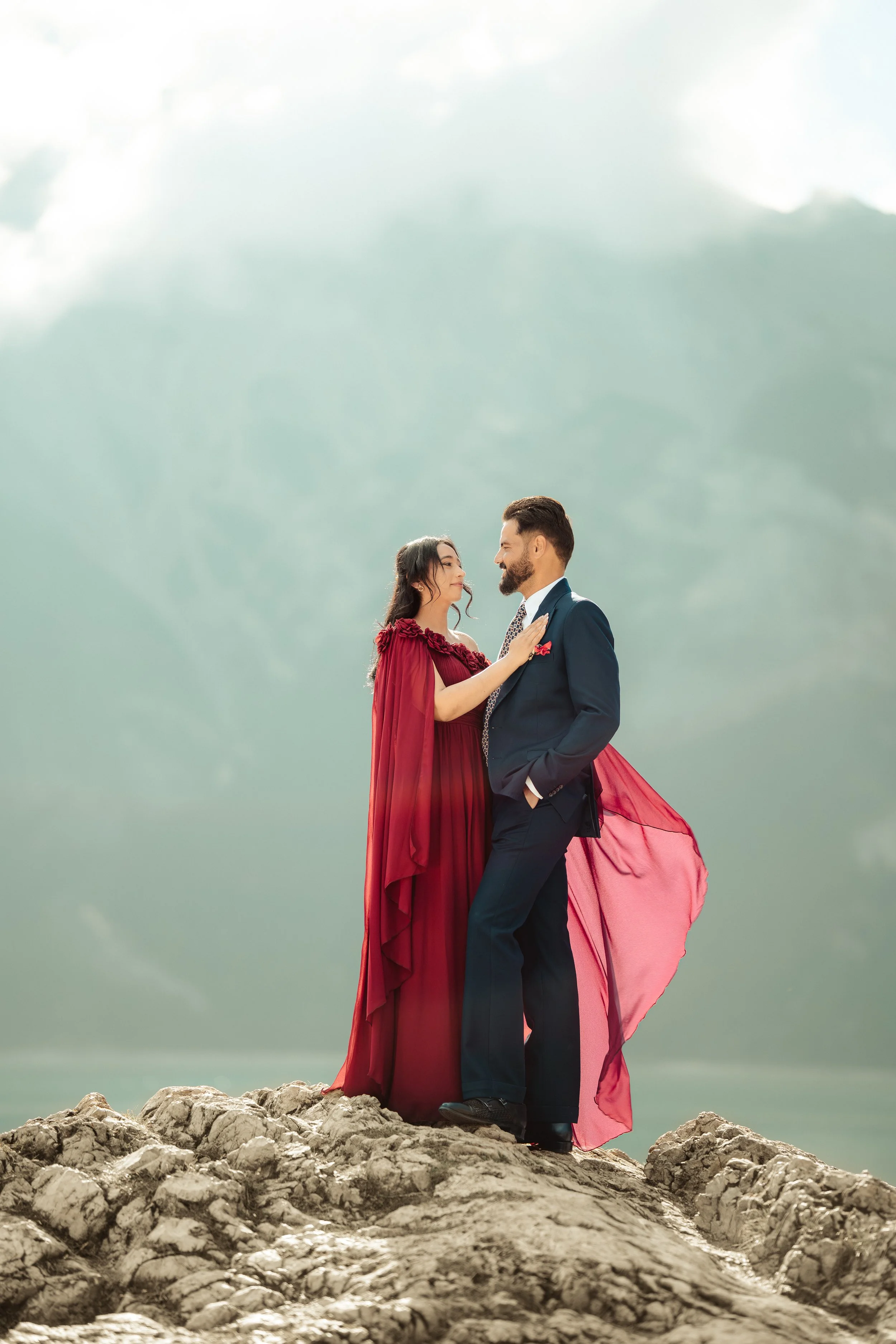 A woman in a red dress and a man in a suit standing on rocks, gazing at each other with a cloudy sky in the background.