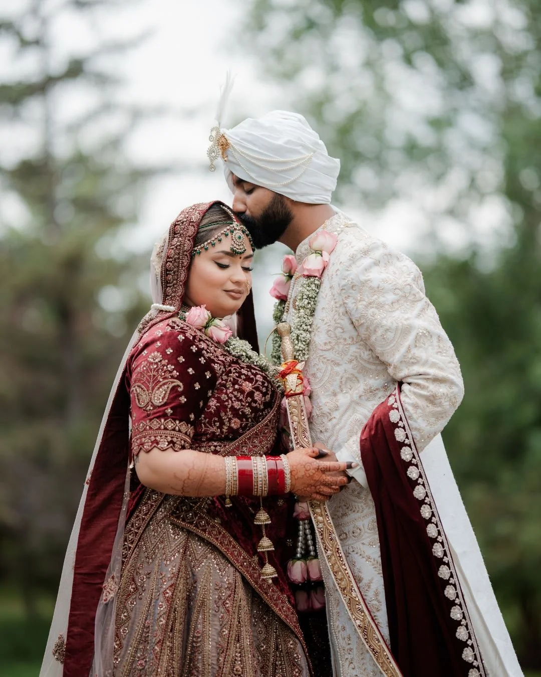 Indian bride and groom in traditional wedding attire, embracing outdoors with the groom kissing the bride's forehead.