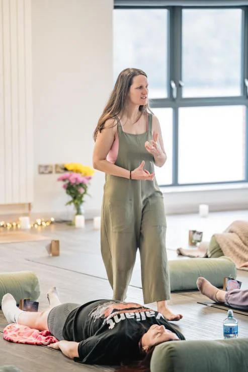 A woman standing and speaking passionately in a large room with large windows, while others are sitting or lying on the floor around her practicing yoga.