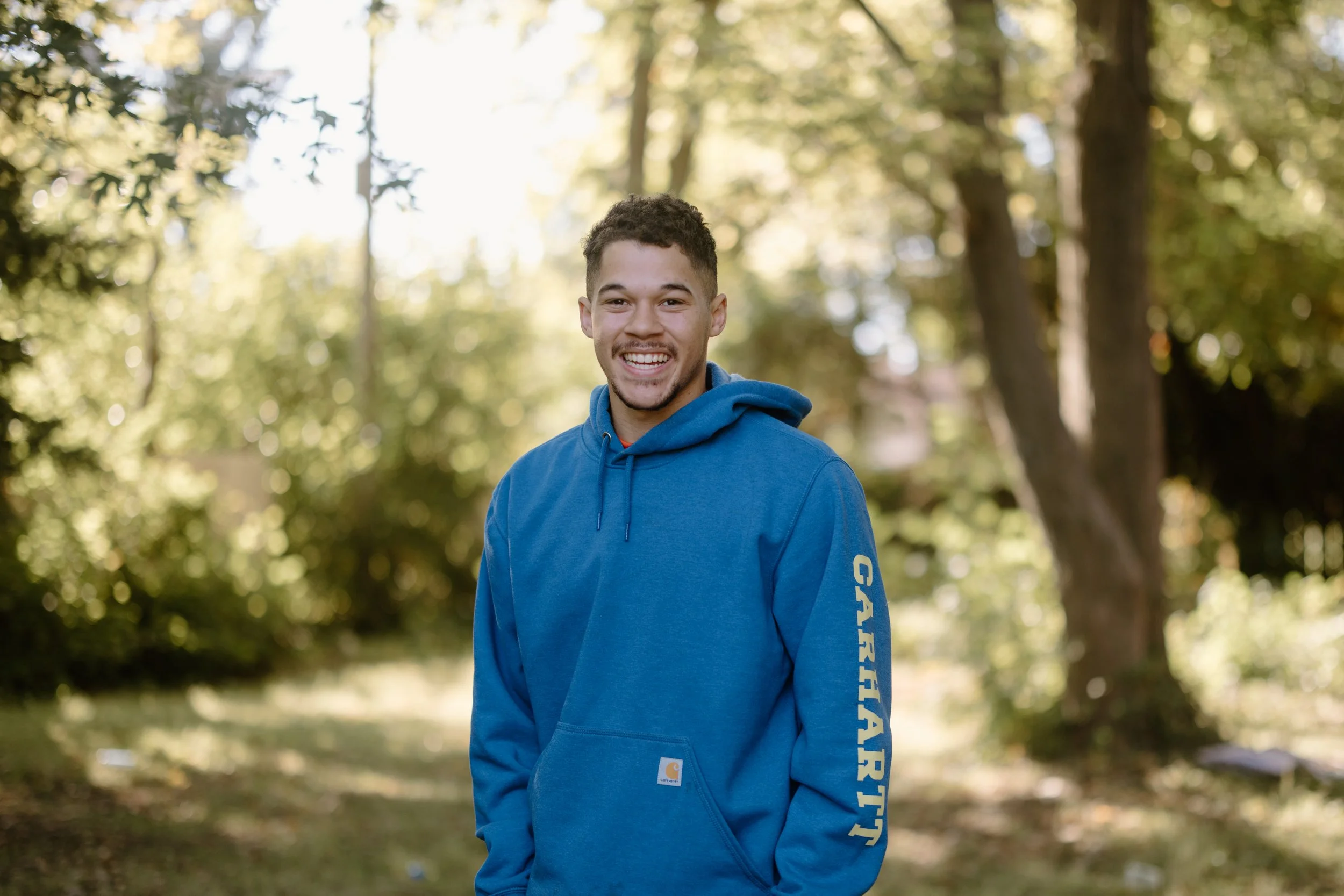 A young man smiling outdoors wearing a blue Carhartt hoodie in a park with trees and sunlight in the background.