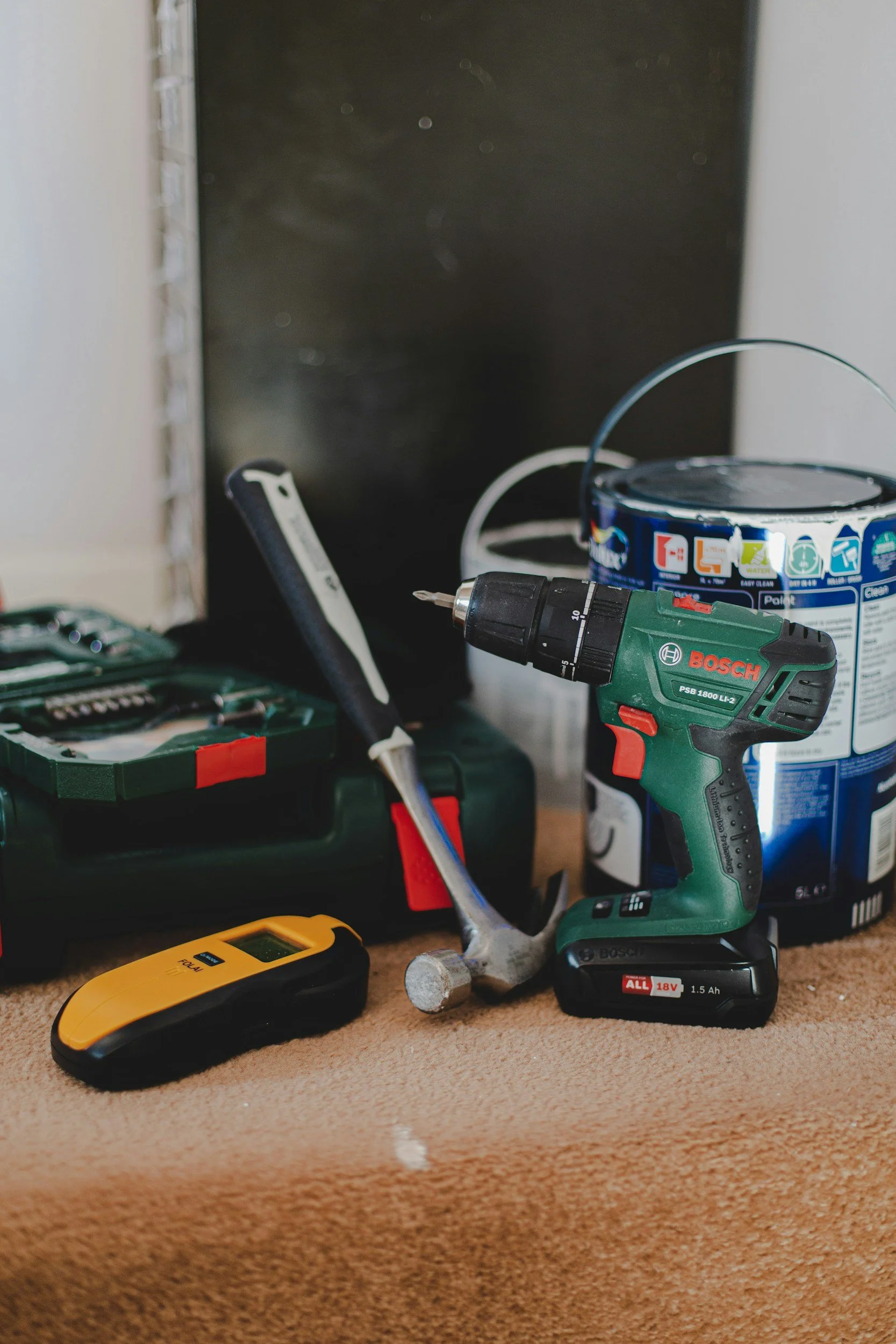 Tools and materials on a workbench, including a cordless drill, a digital device, a small hammer, and cans of paint or stain, with a blackboard or dark background.