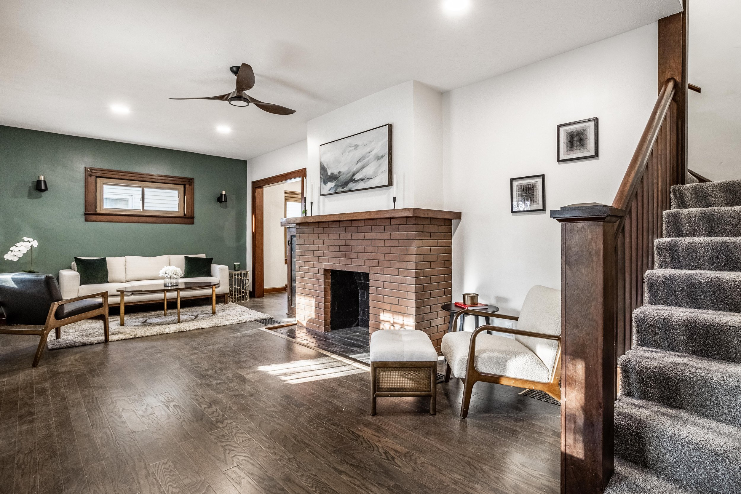 Living room with a brick fireplace, white walls, dark hardwood flooring, and a staircase with grey carpet. Furniture includes a white sofa, black armchair, wooden chair with white cushion, and a beige armchair with a small dark side table. Decor includes framed artwork, a ceiling fan, small wall lights, and a window with wooden trim.