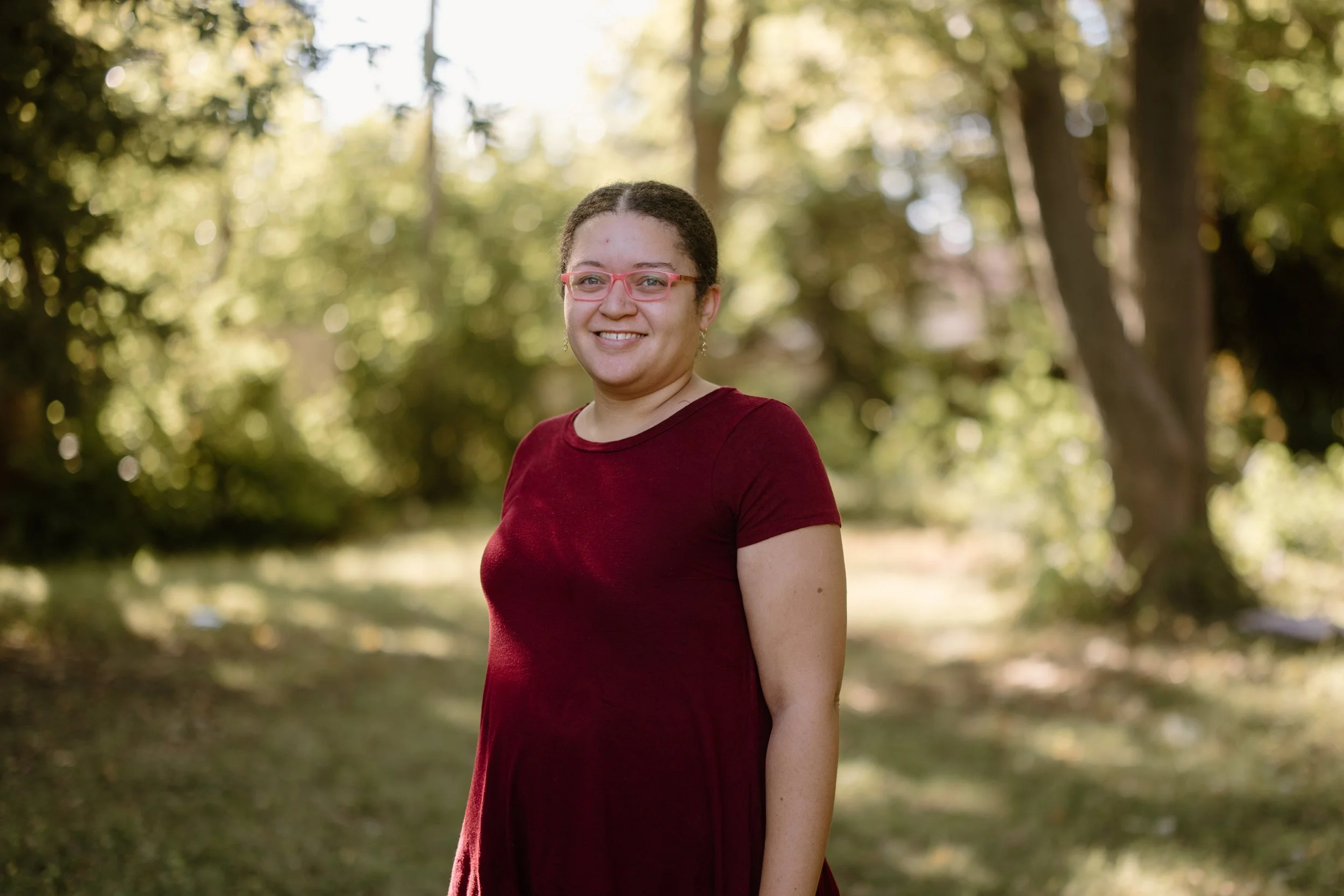 A woman in a red dress and pink glasses standing outdoors with trees and sunlight in the background.