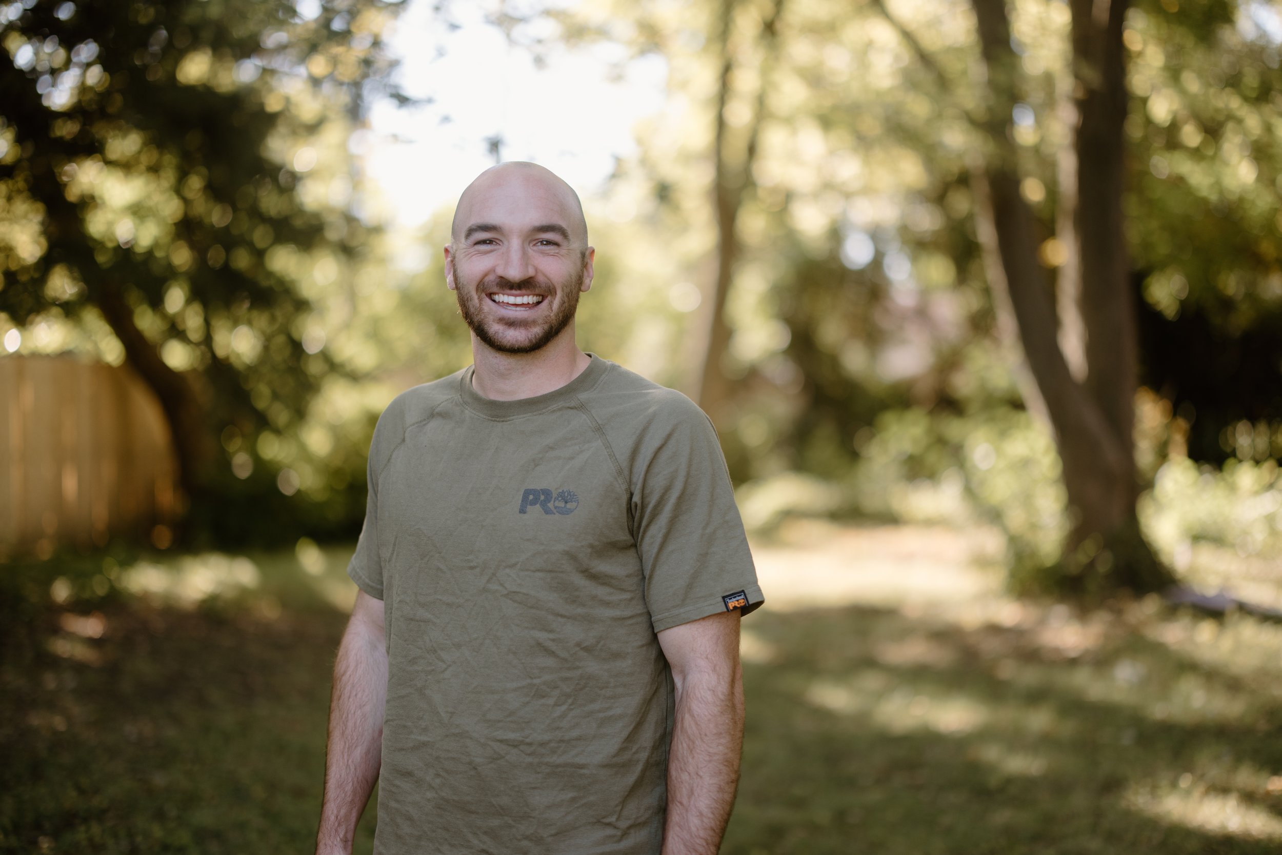 A man with a shaved head and beard smiling outdoors in a wooded area with green trees and sunlight.