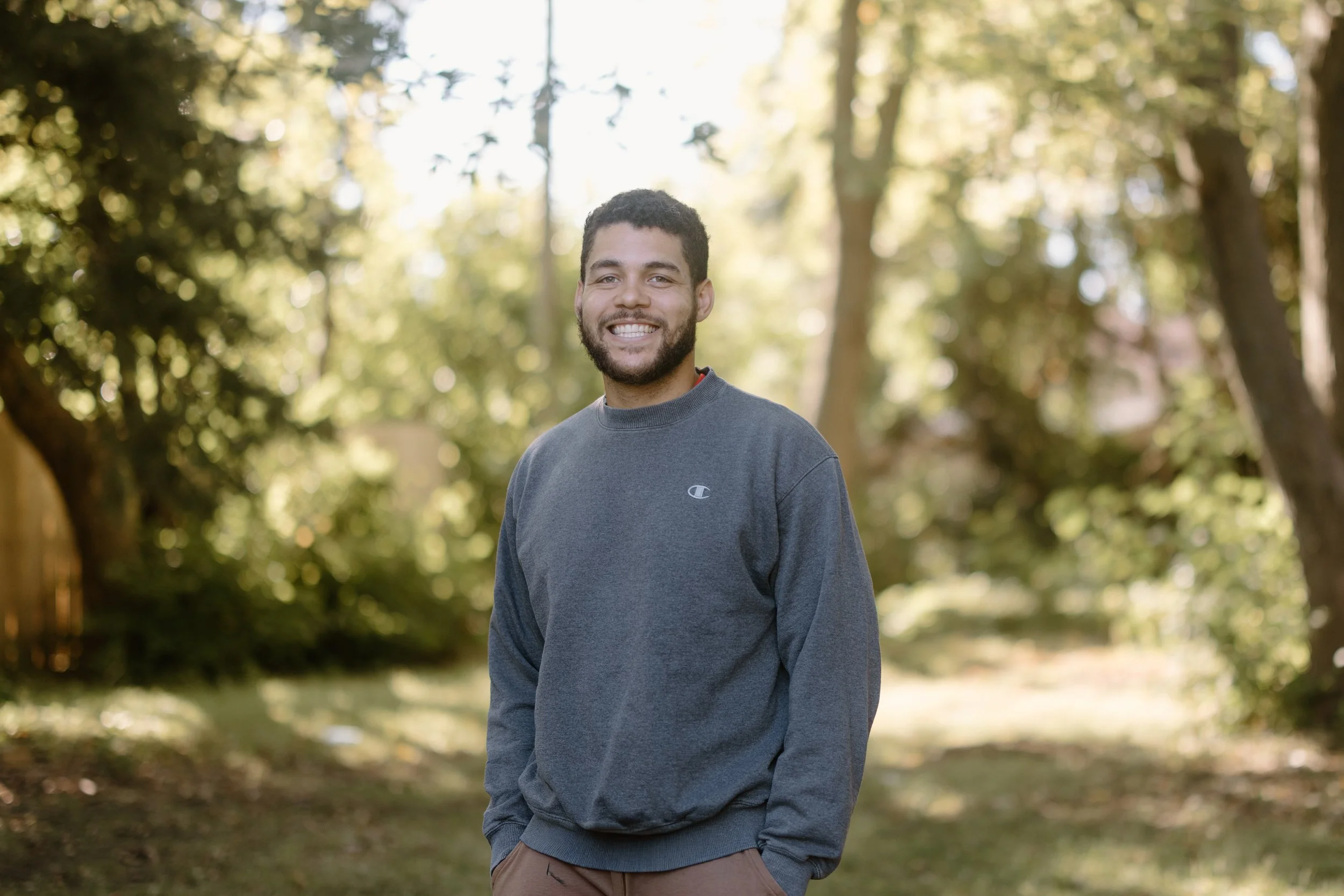 A man smiling outdoors, wearing a gray sweatshirt, standing in a park with trees and sunlight in the background.