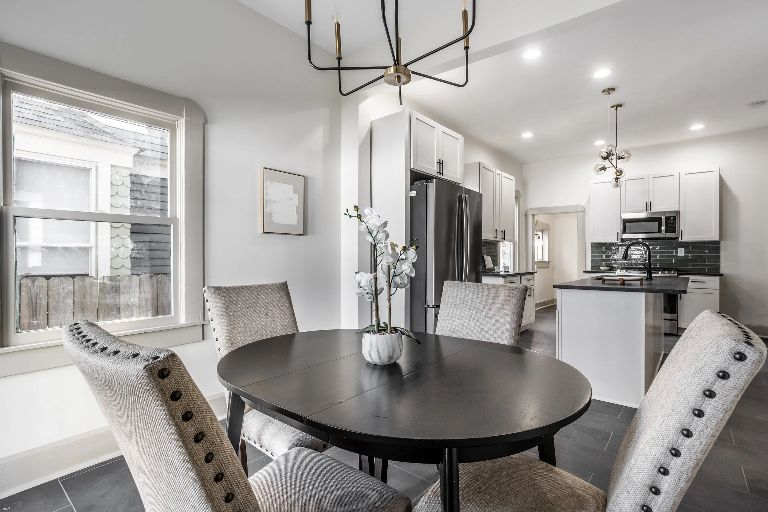 A modern kitchen and dining area with a round black table and four beige upholstered chairs with nailhead trim, a vase with white orchids on the table, large window, white cabinets, stainless steel refrigerator, black backsplash, microwave, and pendant lights.
