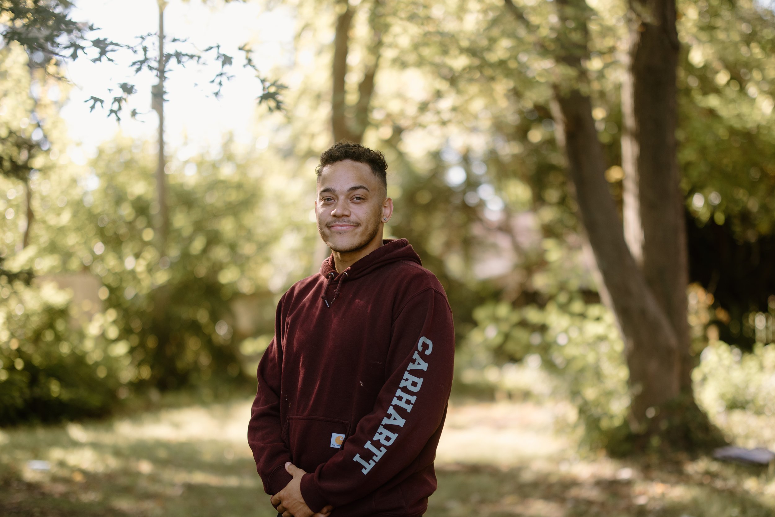 A young man with short dark hair and a slight beard smiling outdoors in a park with trees and sunlight filtering through the leaves. He is wearing a maroon hoodie with the word 'CARHARTT' on the sleeve.