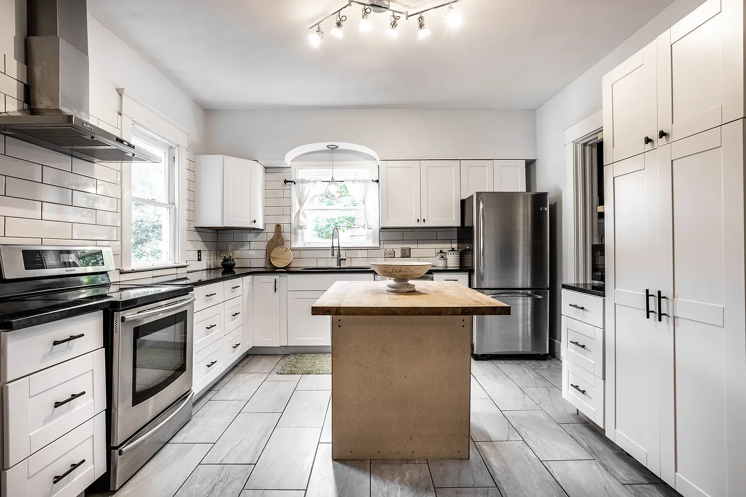 Modern kitchen with white cabinets, stainless steel appliances, a wooden island, black countertops, and a window with white curtains.