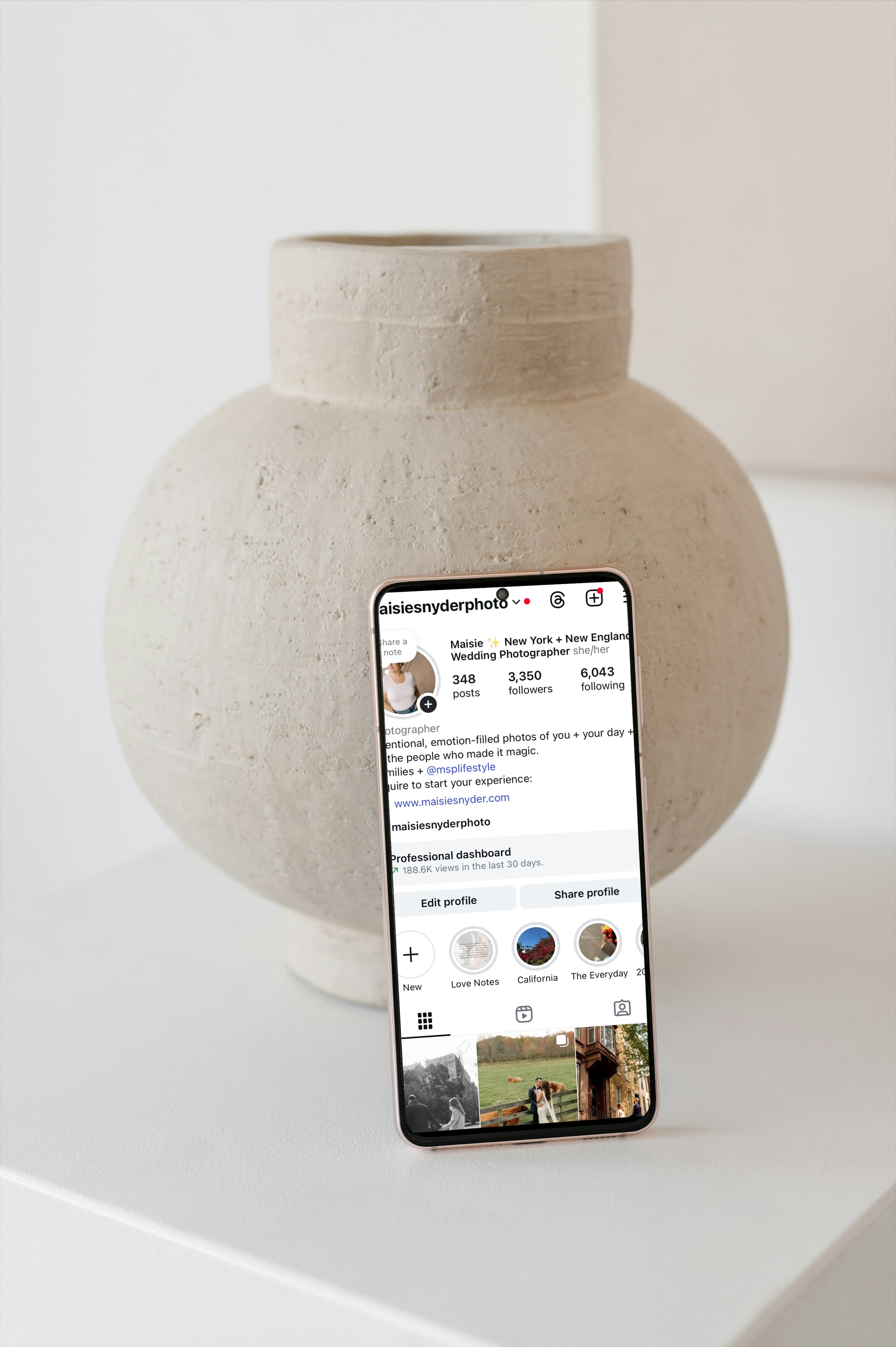 A phone leaning against a ceramic vase displays the @maisiesnyder photo Instagram profile for a New York wedding photographer.