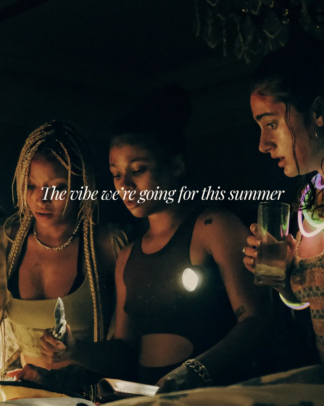Three women huddle together by candlelight and glow sticks at night, with text: 'The vibe we're going for this summer.'