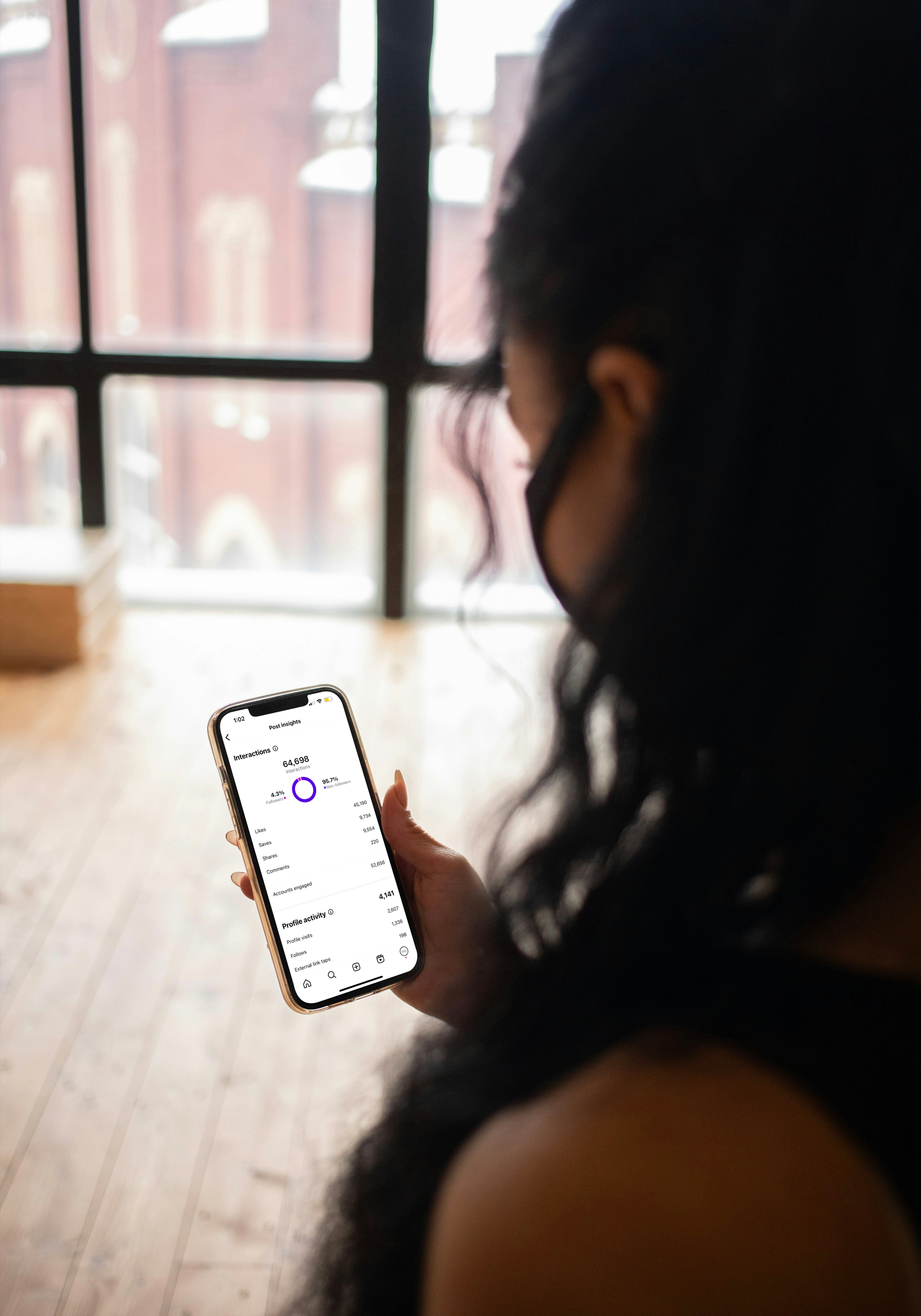 A person reviews Instagram post insights showing 64,698 interactions on a phone, in a sunlit loft studio.