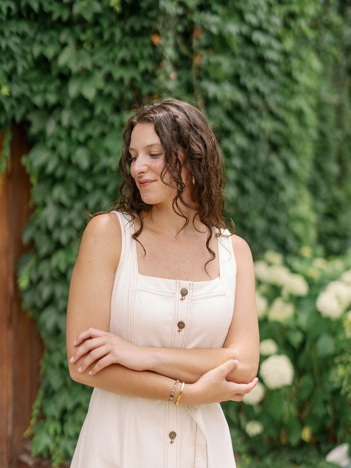 A woman with curly brown hair smiling and looking down, standing outdoors in front of lush green foliage and white flowers, wearing a sleeveless beige dress with buttons.