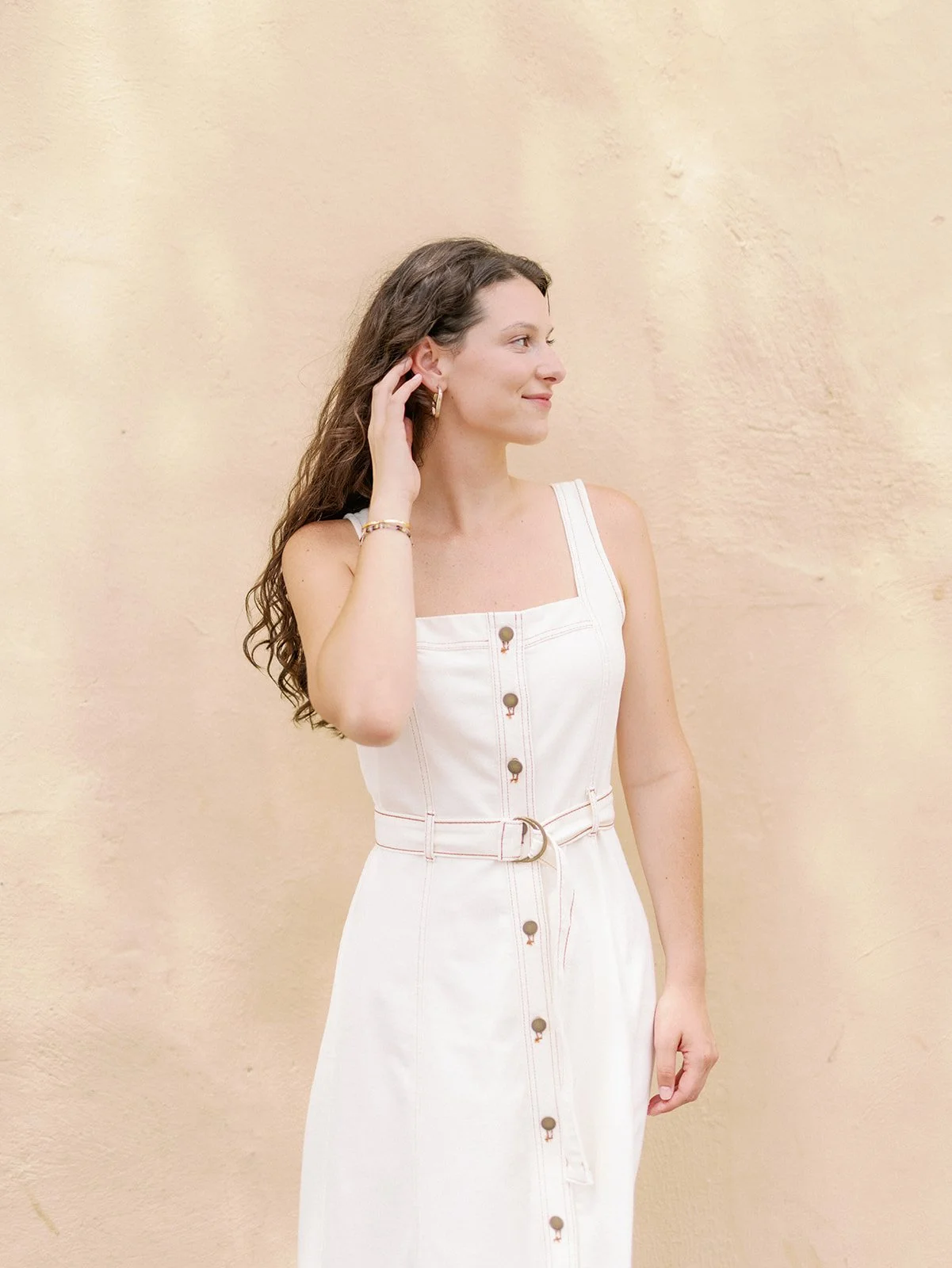 A woman with long, curly brown hair wearing a sleeveless white dress with buttons and a belt, standing against a beige wall, looking to her right and touching her ear.