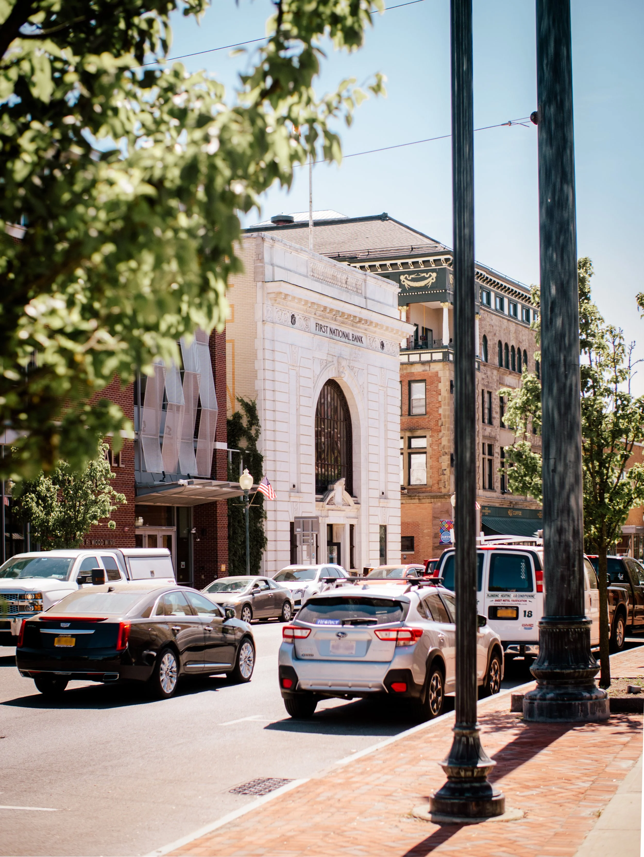Street view with parked cars, trees, vintage lamp posts, and a historic bank building with the sign 'First National Bank' on the facade.