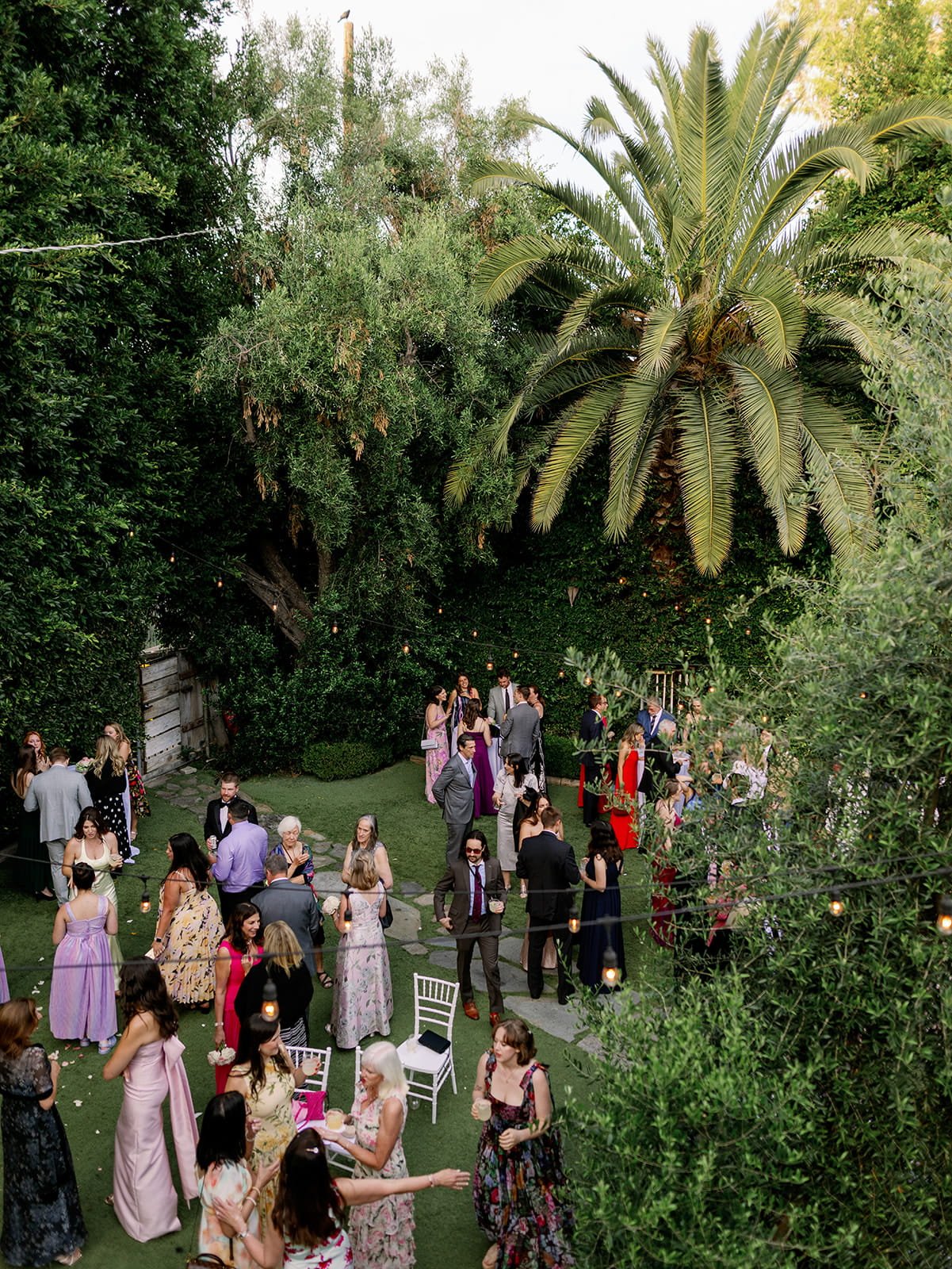 An aerial view of a Los Angeles wedding with guests mingling on a lush garden lawn strung with warm lights, surrounded by palm trees.