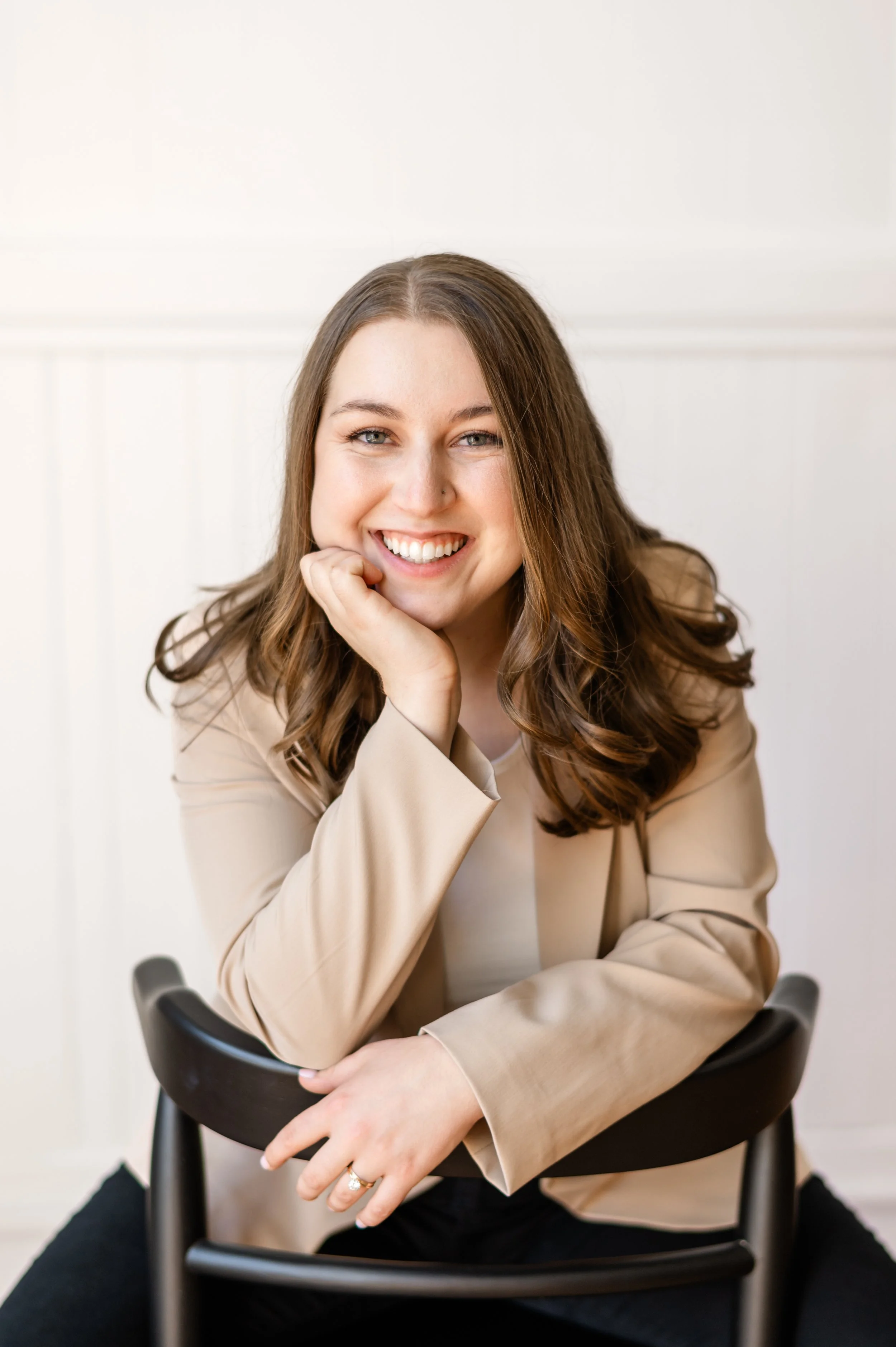 Rebecca Loomis, NY Capital Region brand photographer, leans on a chair smiling warmly in a bright white studio.