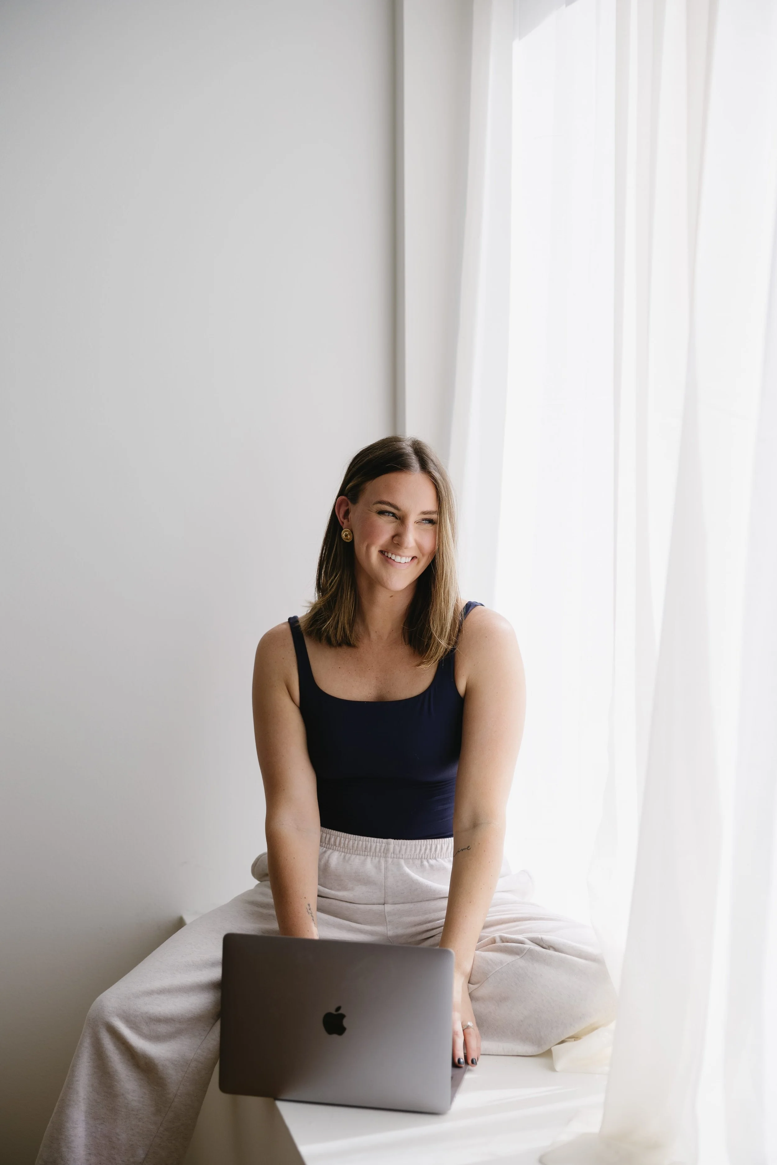 Cassie of Wanders Studio in Glens Falls, NY, with shoulder-length brown hair, wearing a black tank top and beige pants, is sitting on a white couch, smiling and looking at a laptop in front of her.