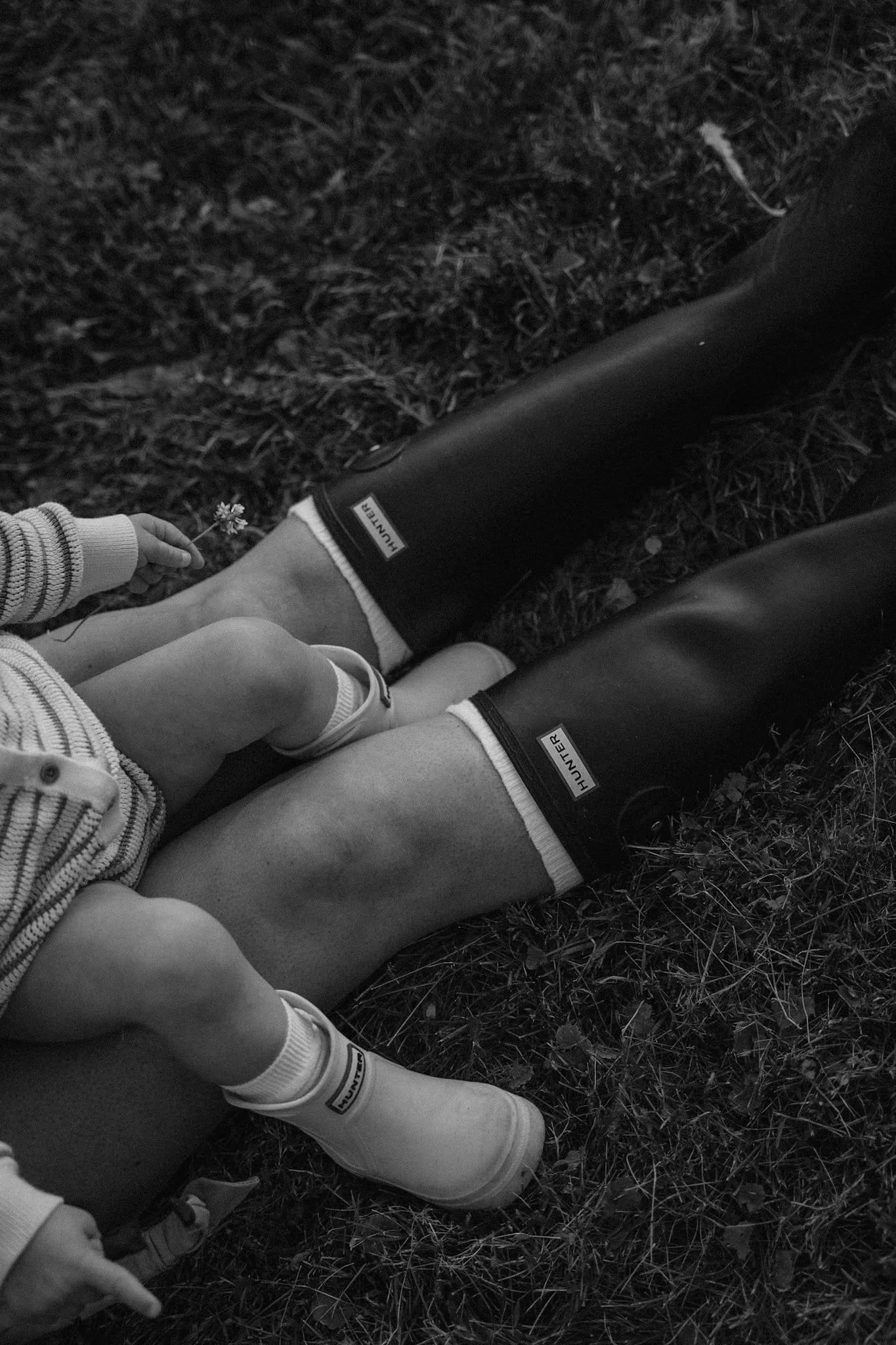 A black-and-white detail shot of a mother and child sitting in grass, both wearing matching Hunter rain boots.
