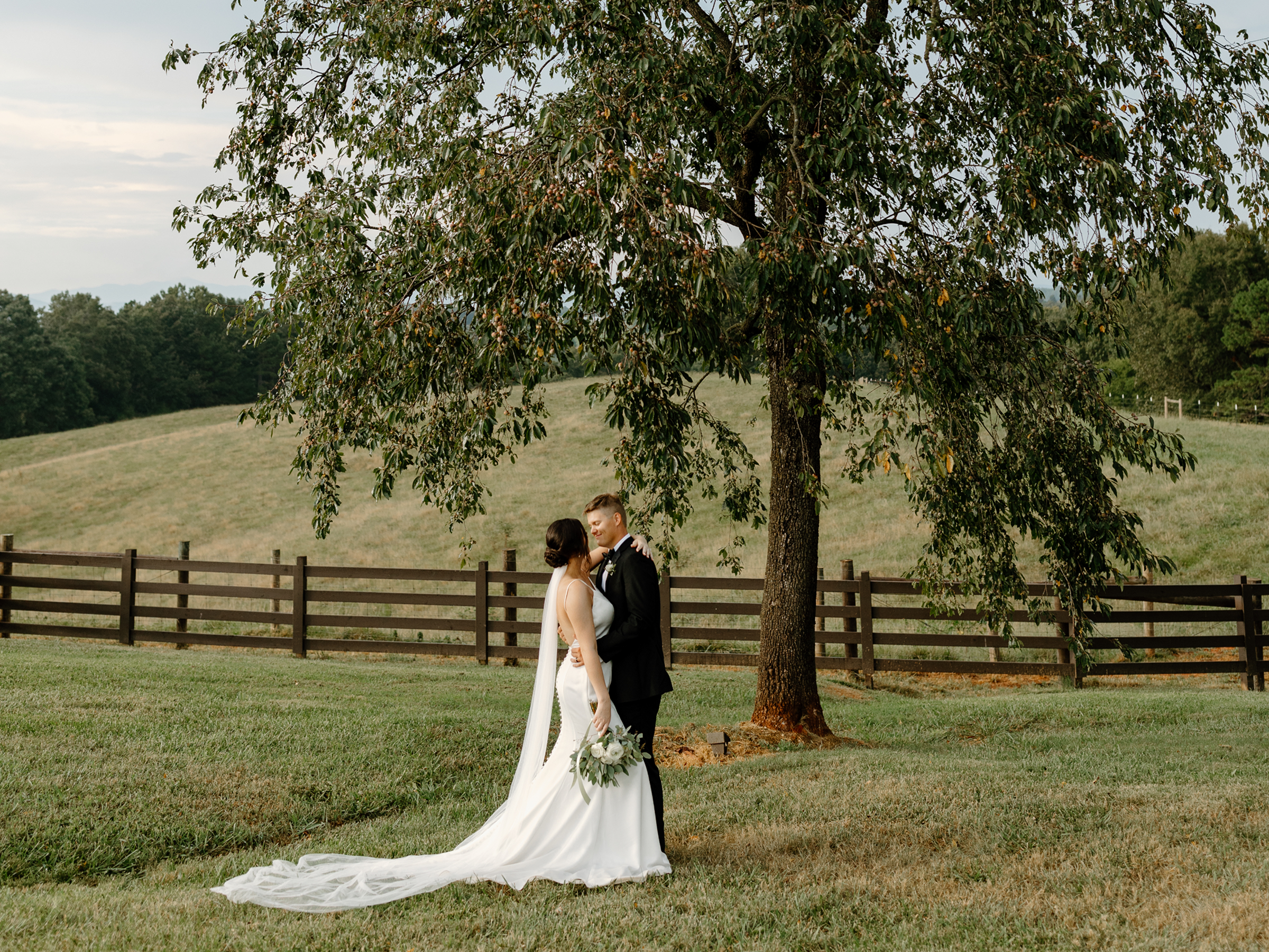 A bride and groom standing under a large tree in a grassy outdoor setting, with a wooden fence behind them. The bride is wearing a white wedding dress and holding a bouquet, and the groom is in a black suit.