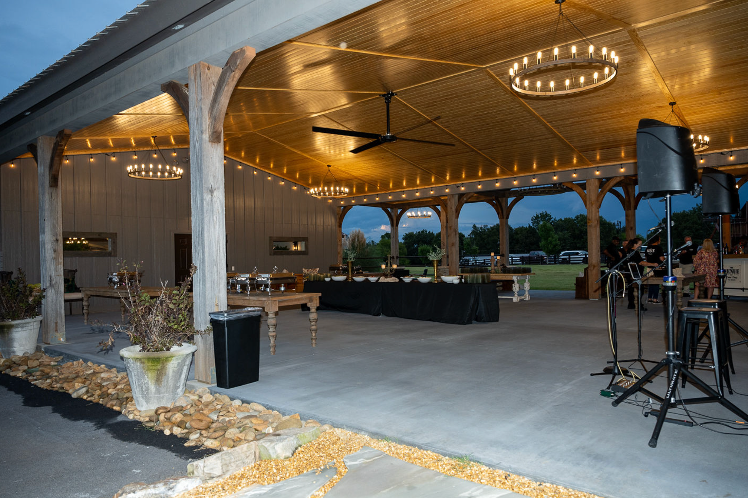 Outdoor event space with a wooden ceiling and chandeliers, tables with decorations, and sound equipment, set up for a celebration or gathering during early evening.