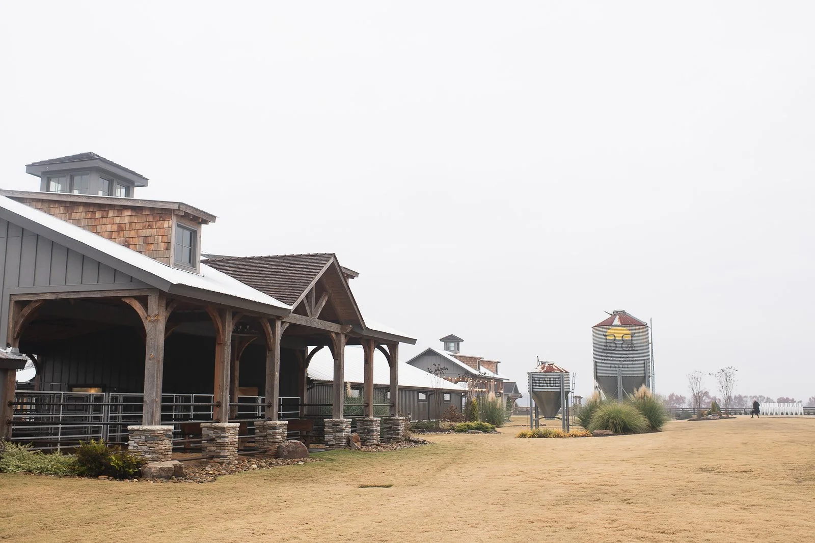 Farm buildings with large grain silos, grassy field, and overcast sky.