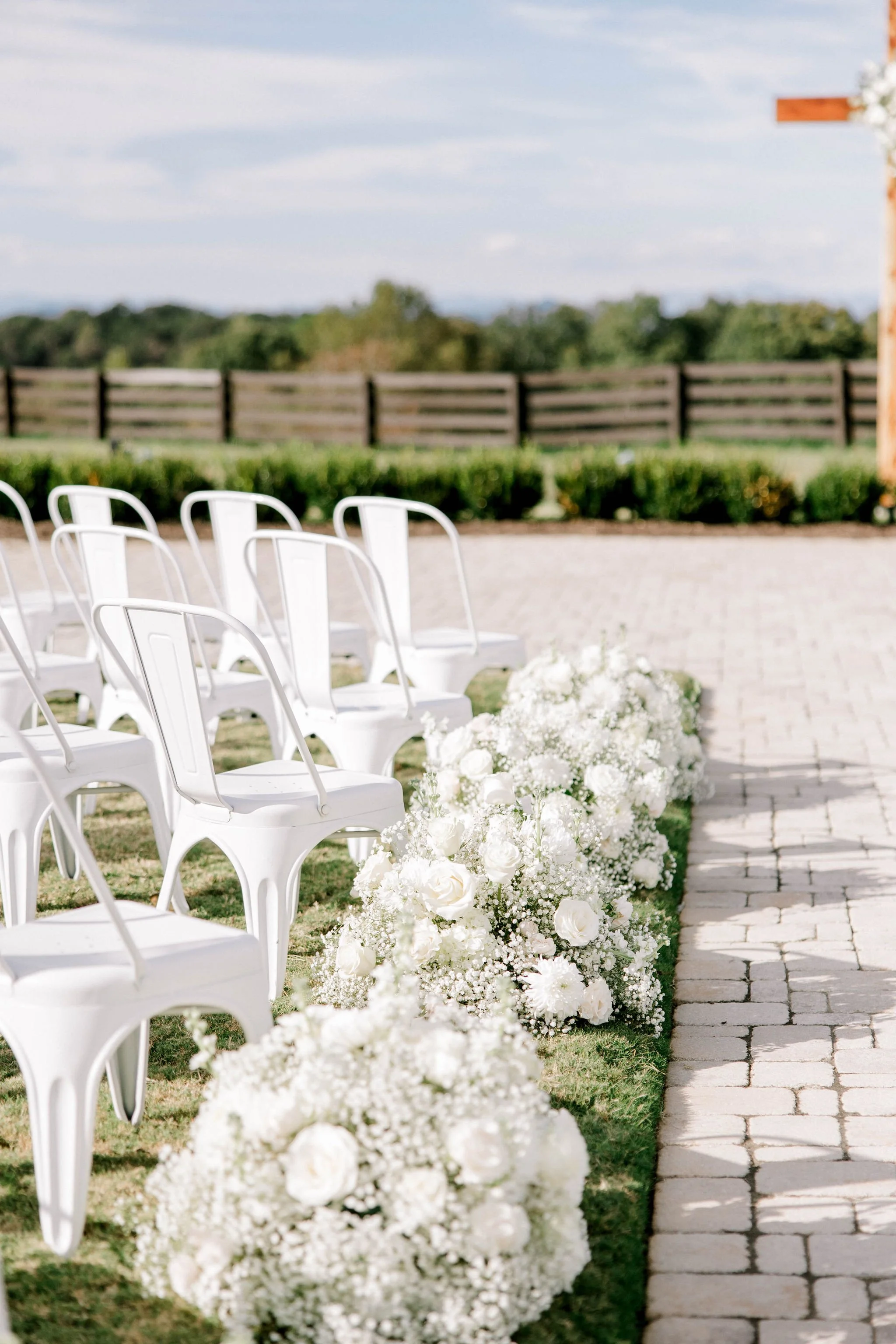 White chairs arranged outdoors for a wedding ceremony, with white floral arrangements along a stone pathway and a wooden fence in the background.