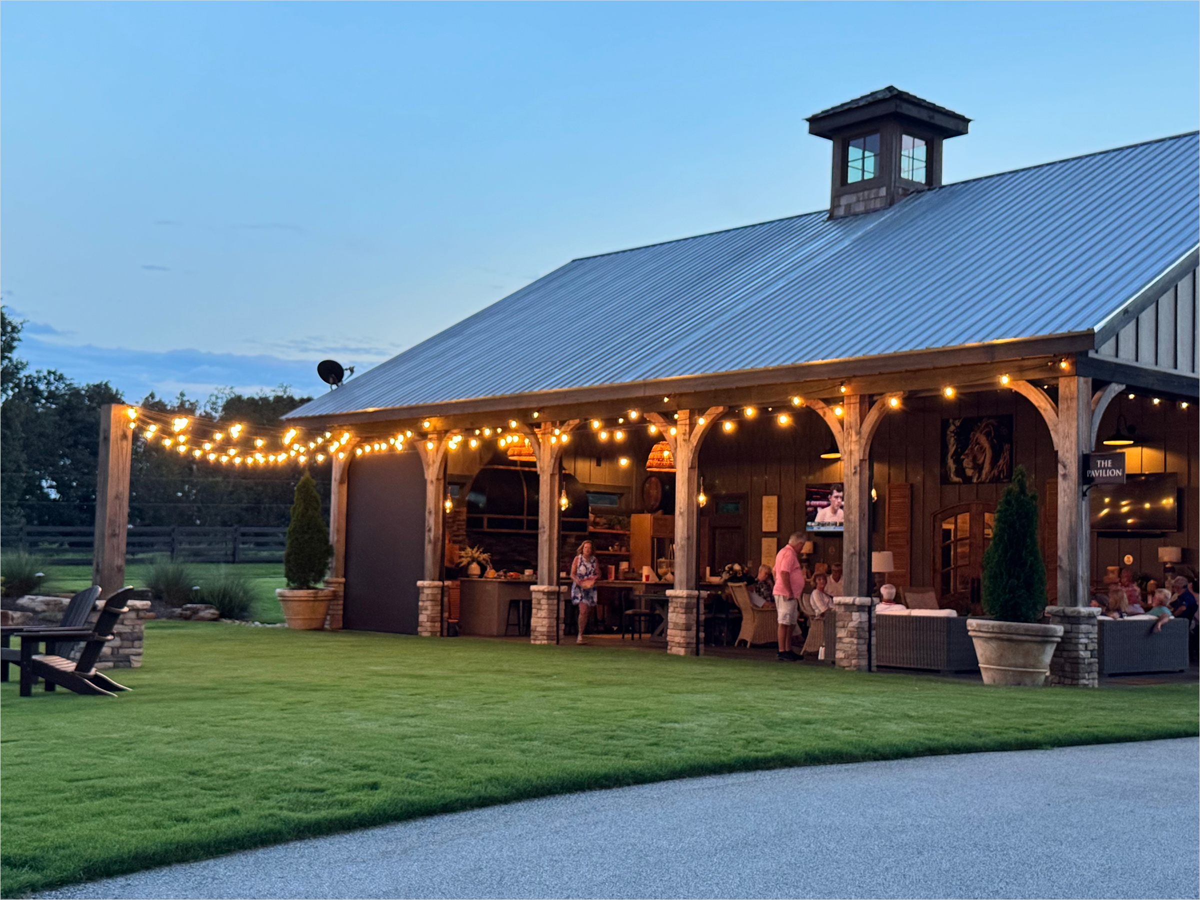 Outdoor evening scene of a rustic building with a metal roof, string lights, and outdoor seating, with people dining inside and outside.