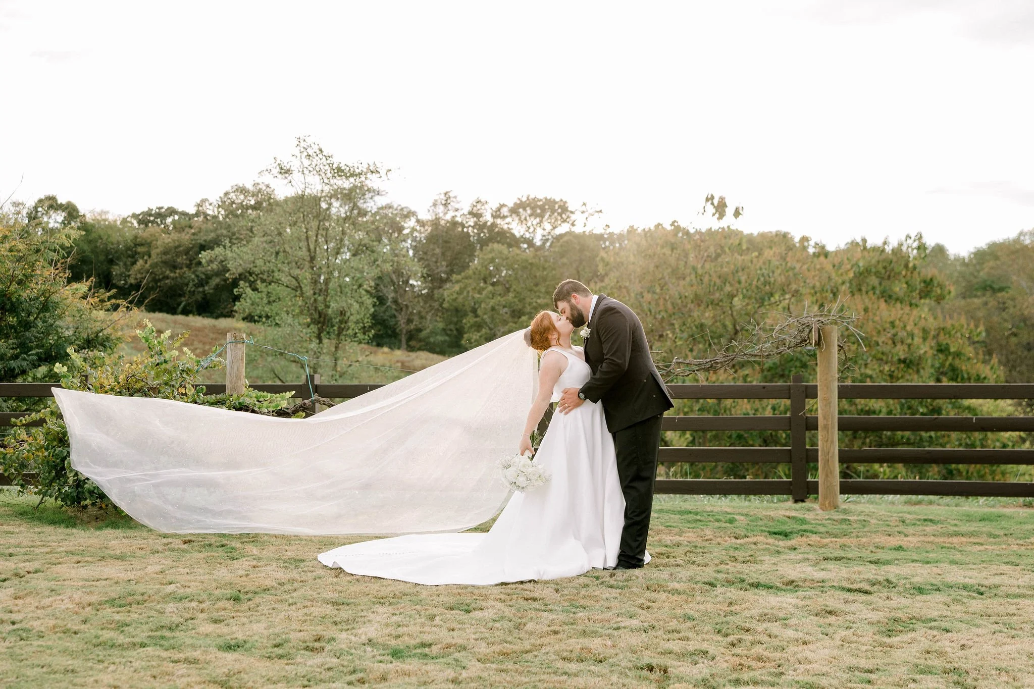 A bride and groom share a kiss outdoors on their wedding day, with the bride in a white gown holding a bouquet and her long veil flowing behind her, and the groom in a black tuxedo, against a background of trees and a wooden fence.