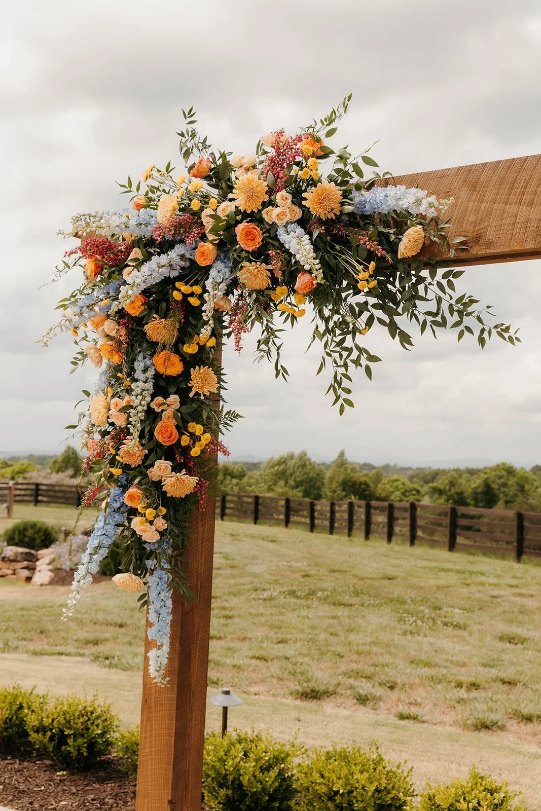 Floral arrangement on a wooden arch at an outdoor wedding, featuring pink, yellow, orange, and blue flowers with greenery, under an overcast sky.
