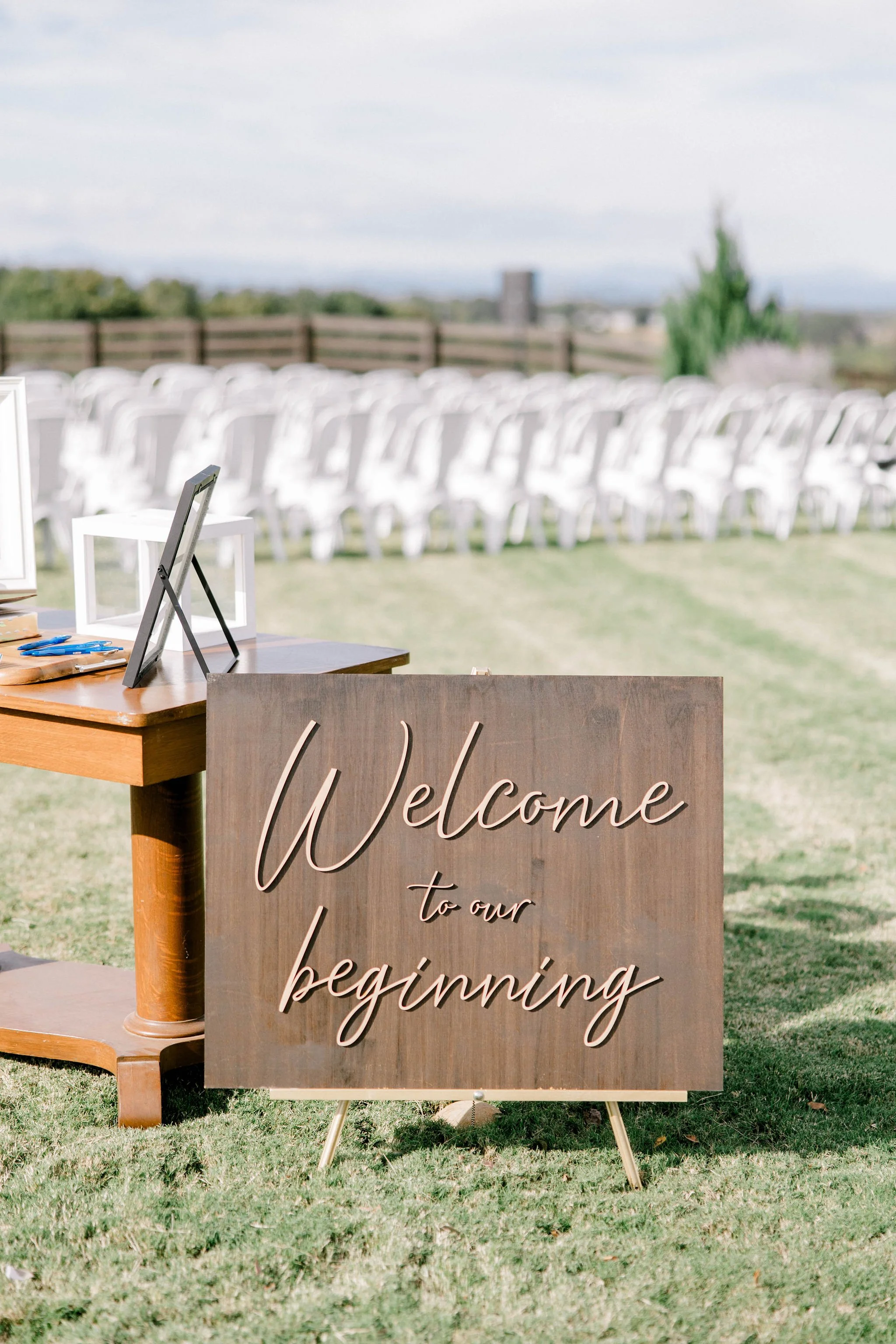 A wooden sign on a lawn reads "Welcome to our beginning" with rows of white chairs in the background, set up outdoors for an event.