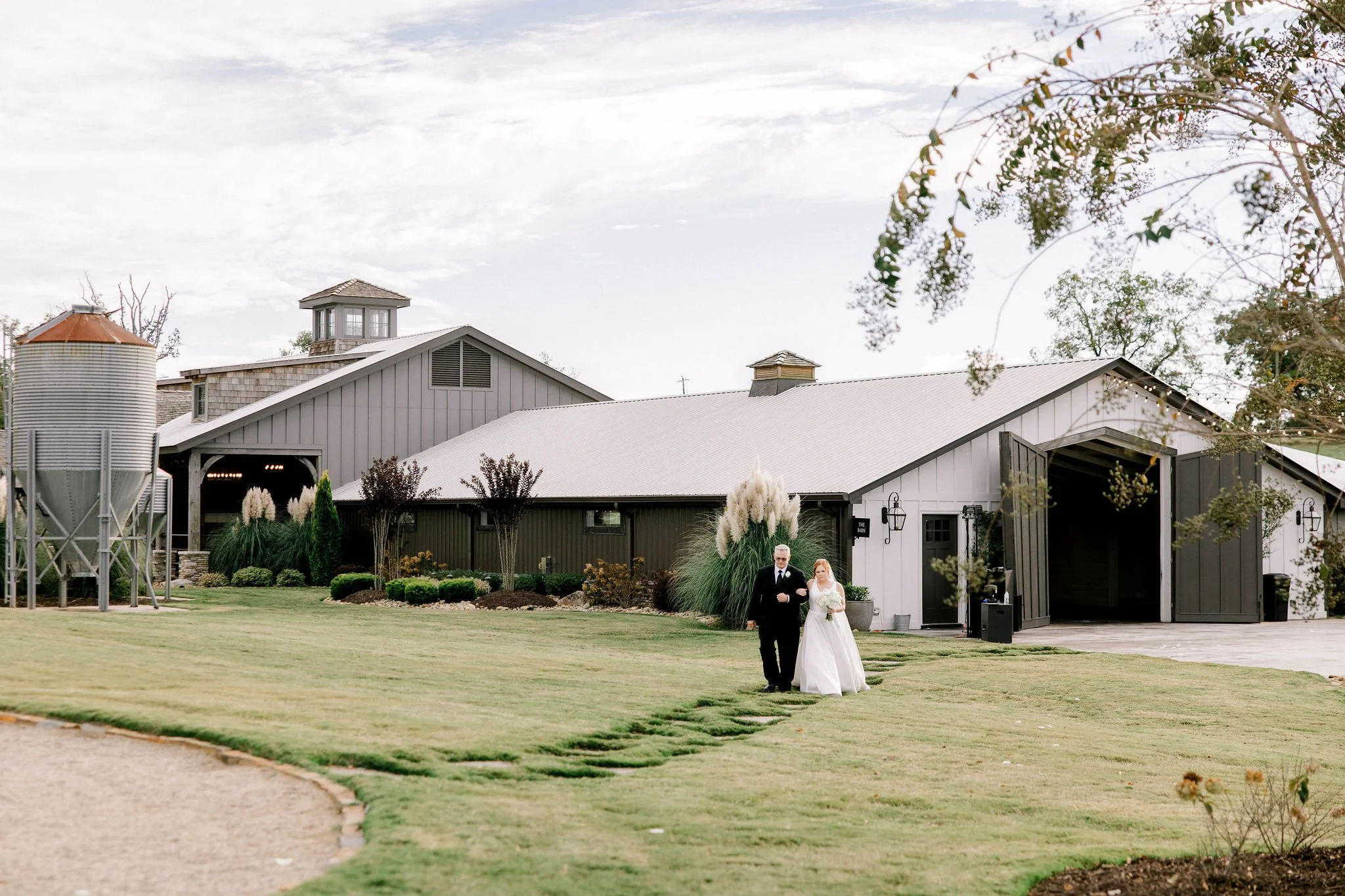 A bride and a groom walking on a grassy lawn outside a rustic barn, with the bride holding a bouquet, amid decorative plants and trees.