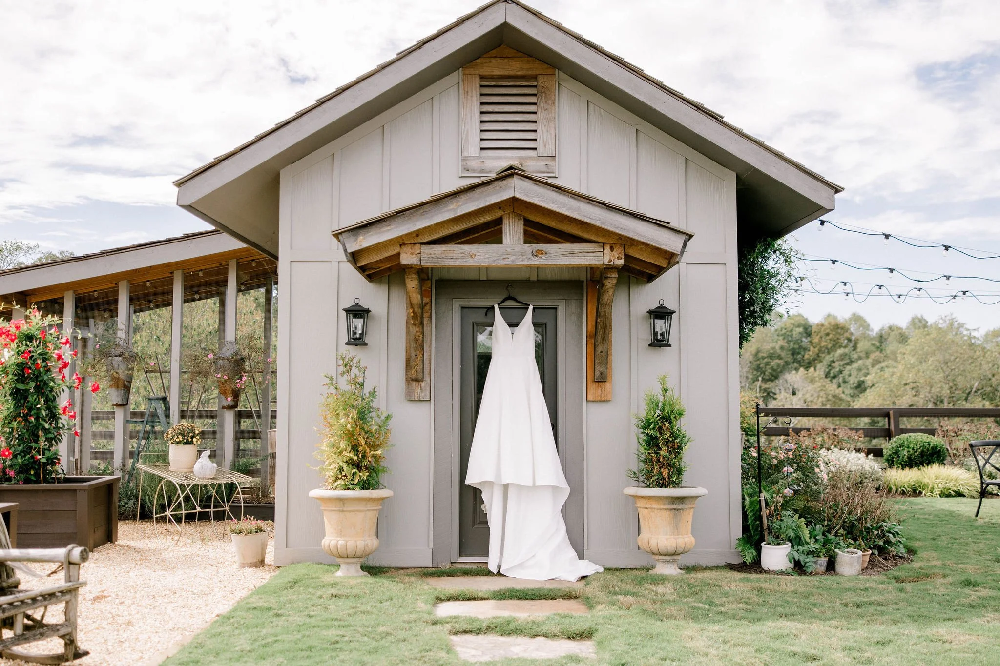 White wedding dress hanging on a black hanger in front of a small barn with outdoor string lights and potted plants.