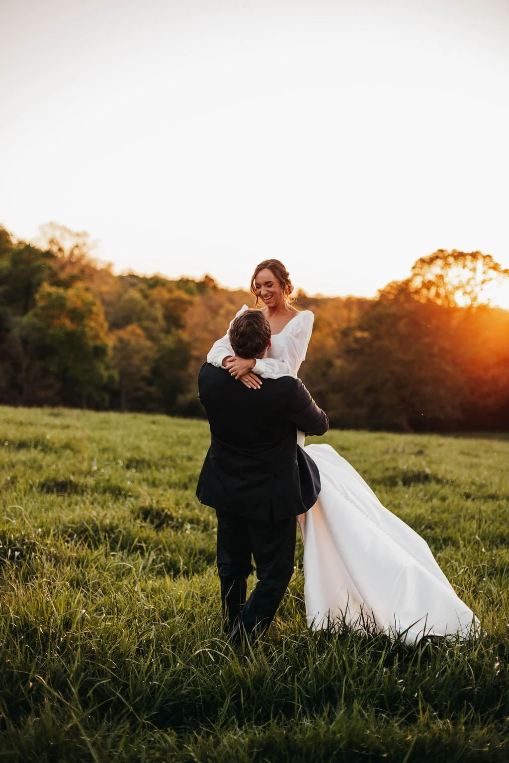 A bride and groom sharing a joyful moment outdoors during sunset, with the bride being lifted by the groom in a grassy field surrounded by trees.