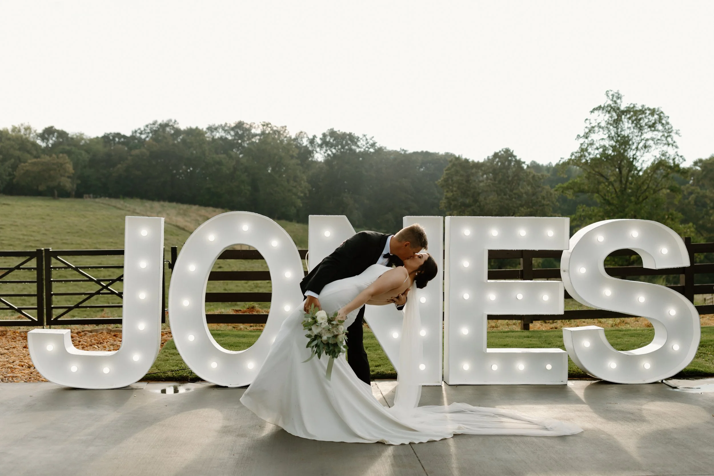 A bride and groom sharing a kiss in front of large illuminated letters spelling 'LOVE' outdoors with a green field and trees in the background.