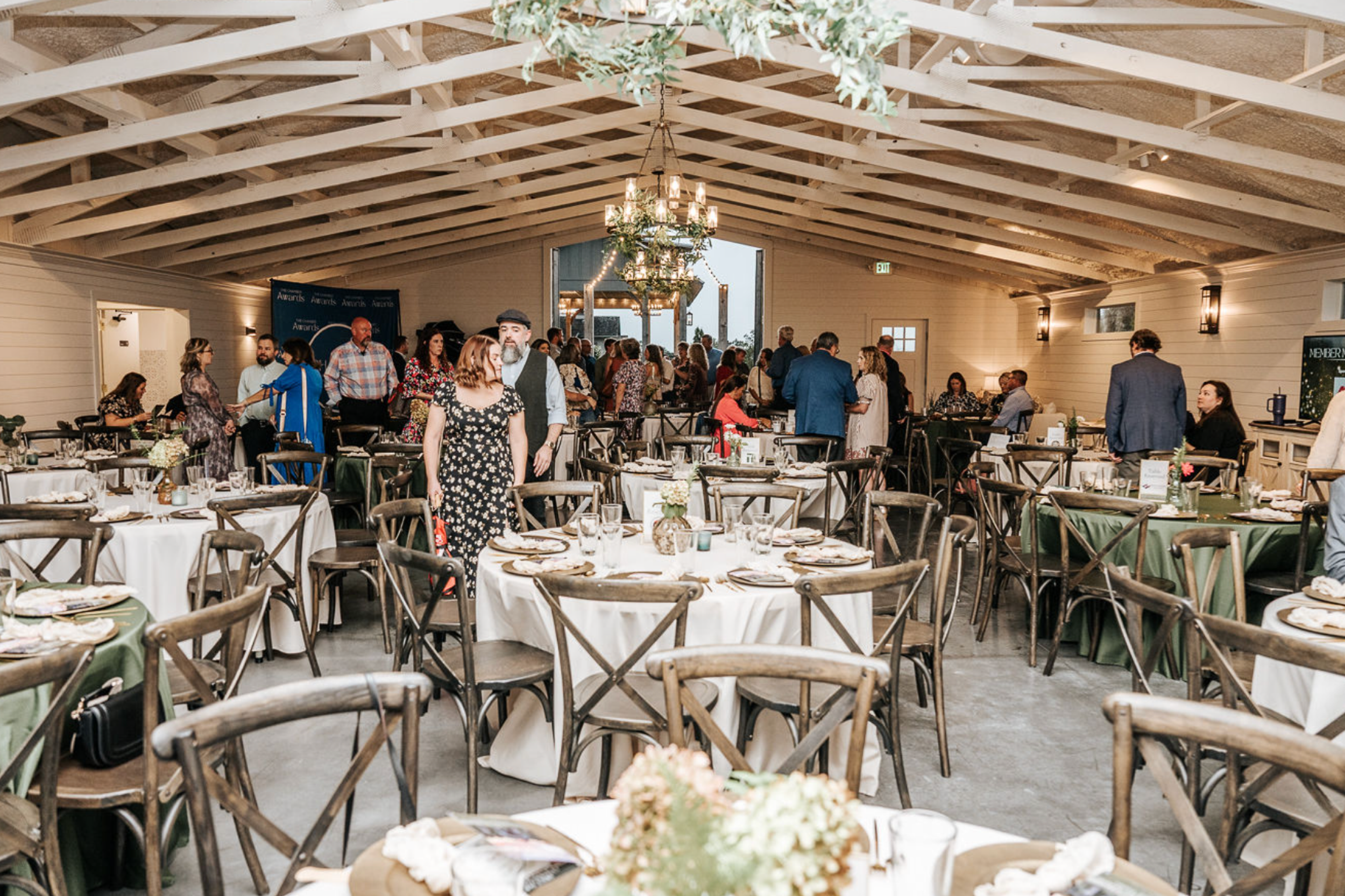 Interior of a rustic-style event space with round tables set for a meal, wooden chairs, and a chandelier, with people mingling and socializing.