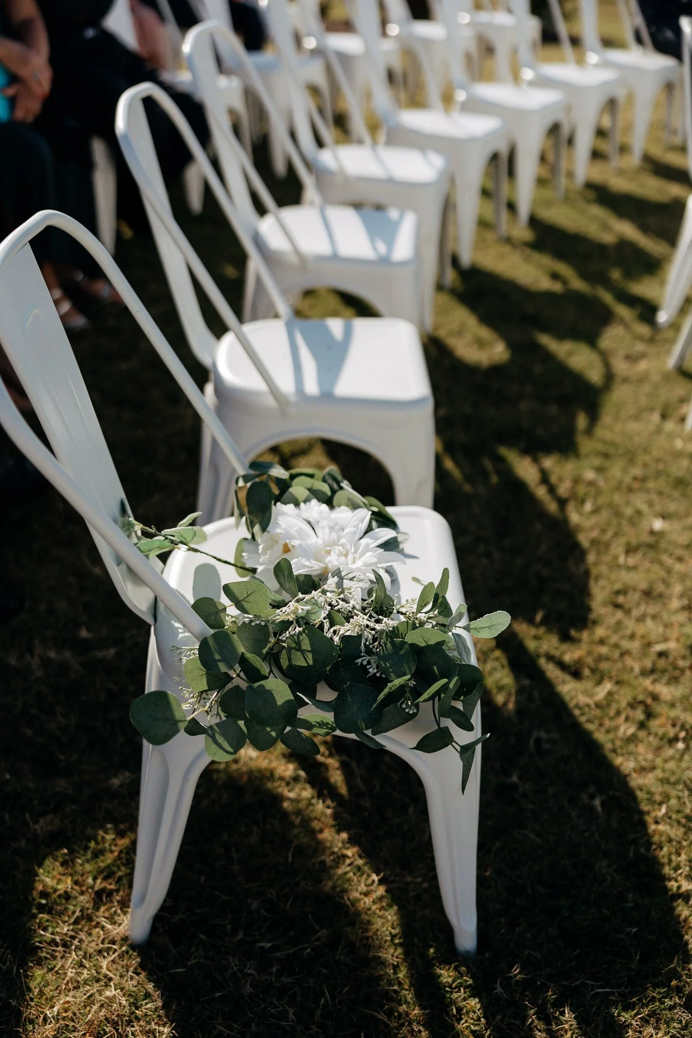 White chairs arranged in a row at an outdoor wedding ceremony with one chair decorated with white flowers and green leaves.