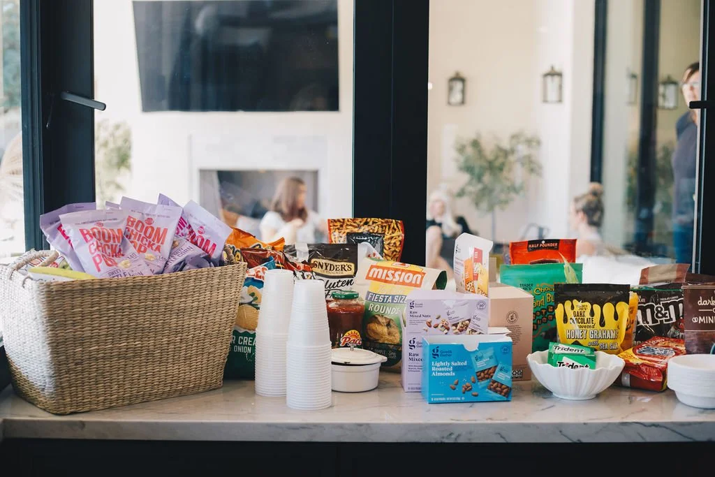 A variety of snack foods and drinks arranged on a kitchen or dining table near a window.