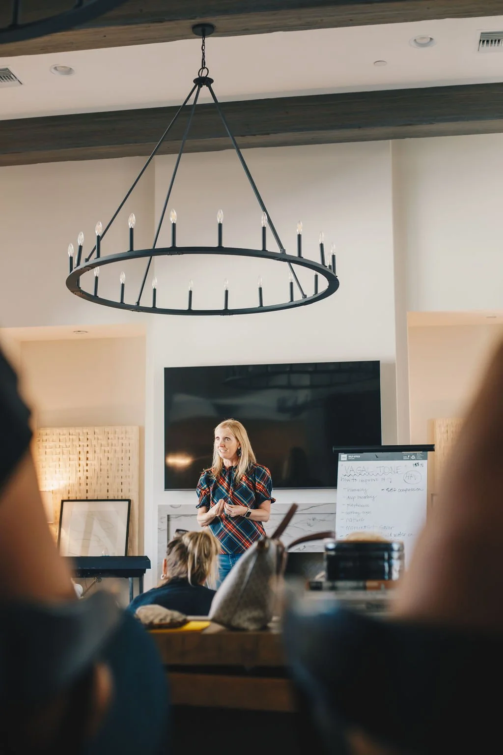 A woman giving a presentation in a conference room with a large chandelier hanging from the ceiling, a black television on the wall, and a whiteboard with notes.