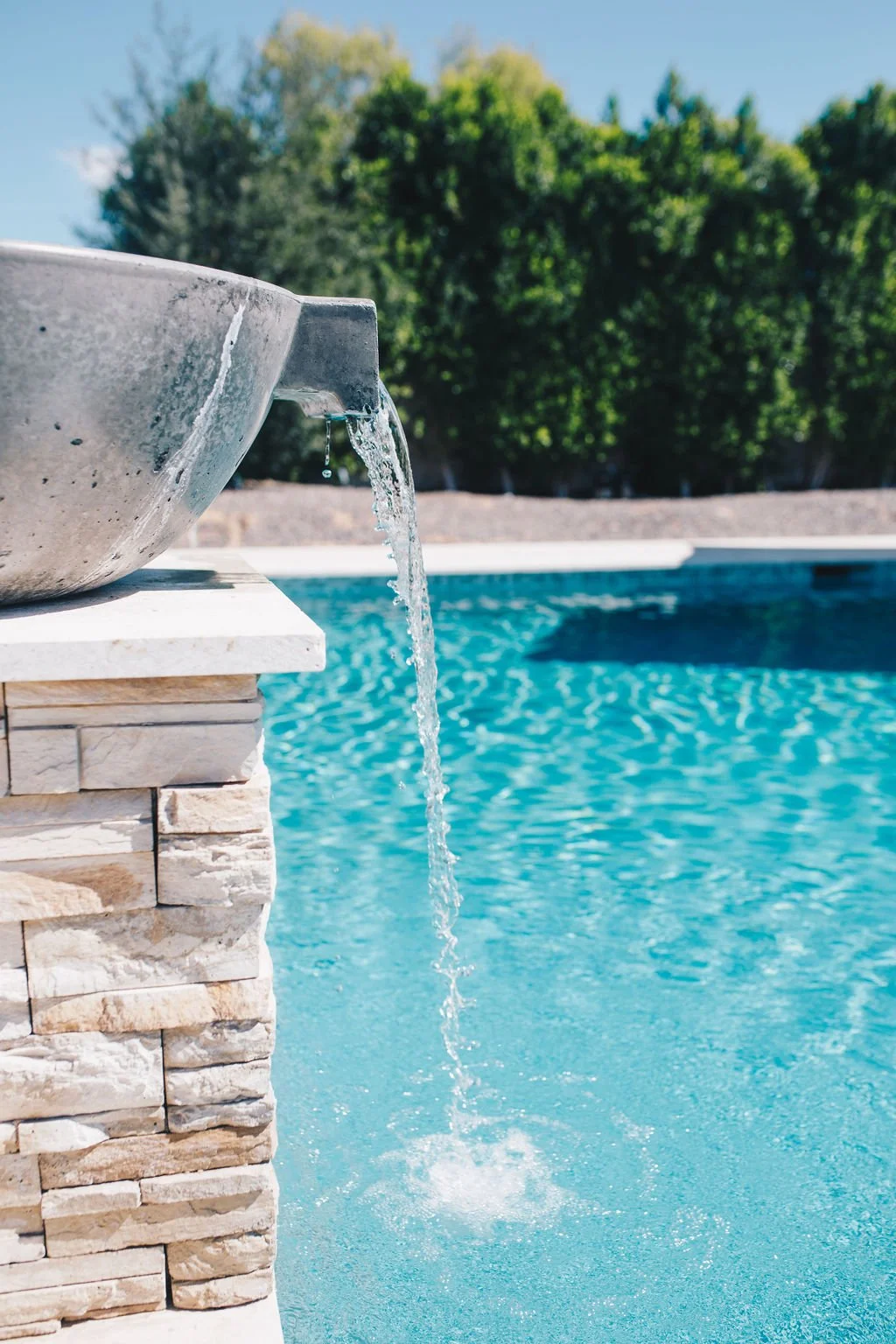 Water flowing from a stone fountain into a swimming pool with a backdrop of green trees under a clear blue sky.