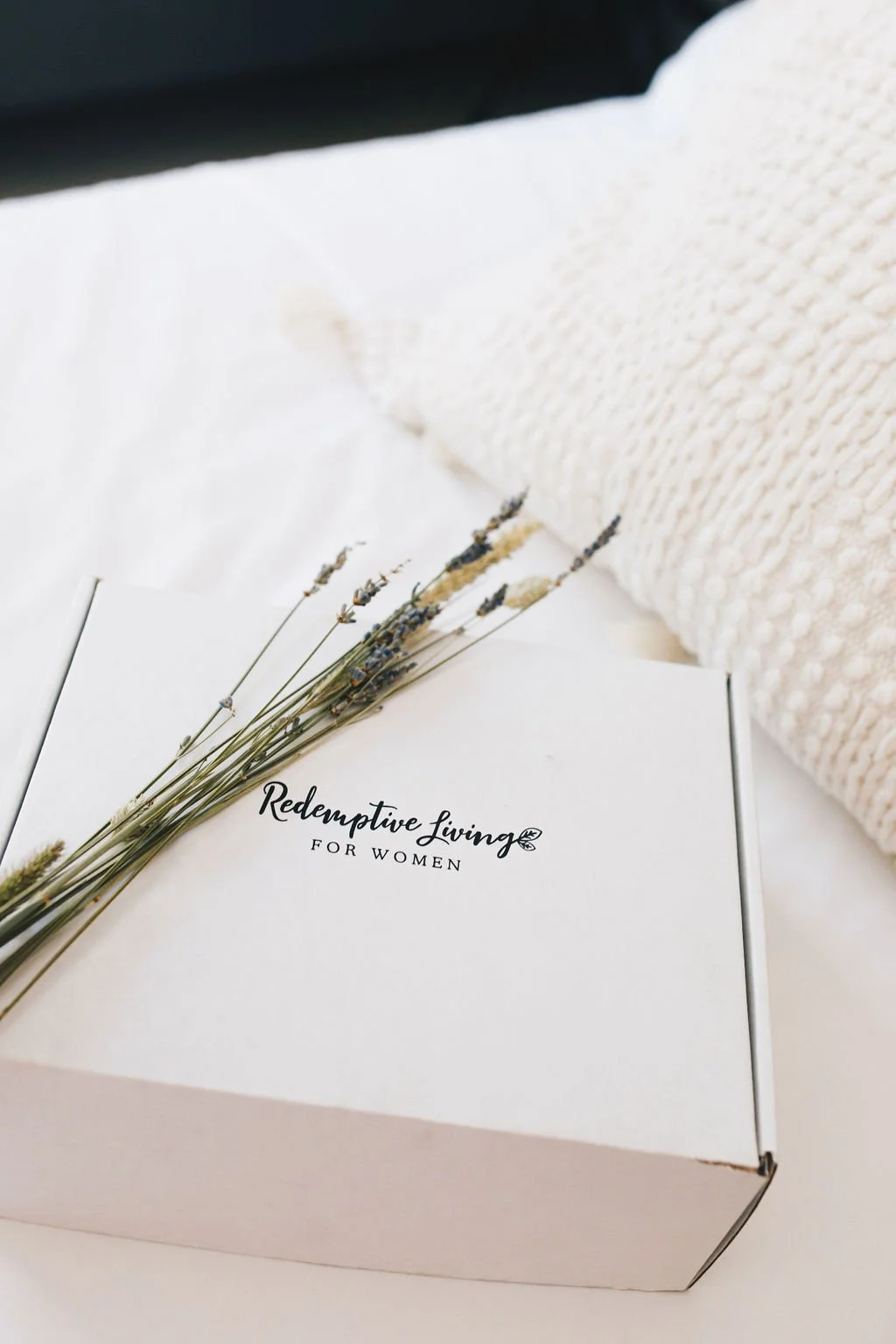 A white box with black text titled 'Redemptive Living for Women' resting on white fabric, with a bunch of dried lavender flowers laid on top, and a cream-colored knitted blanket in the background.