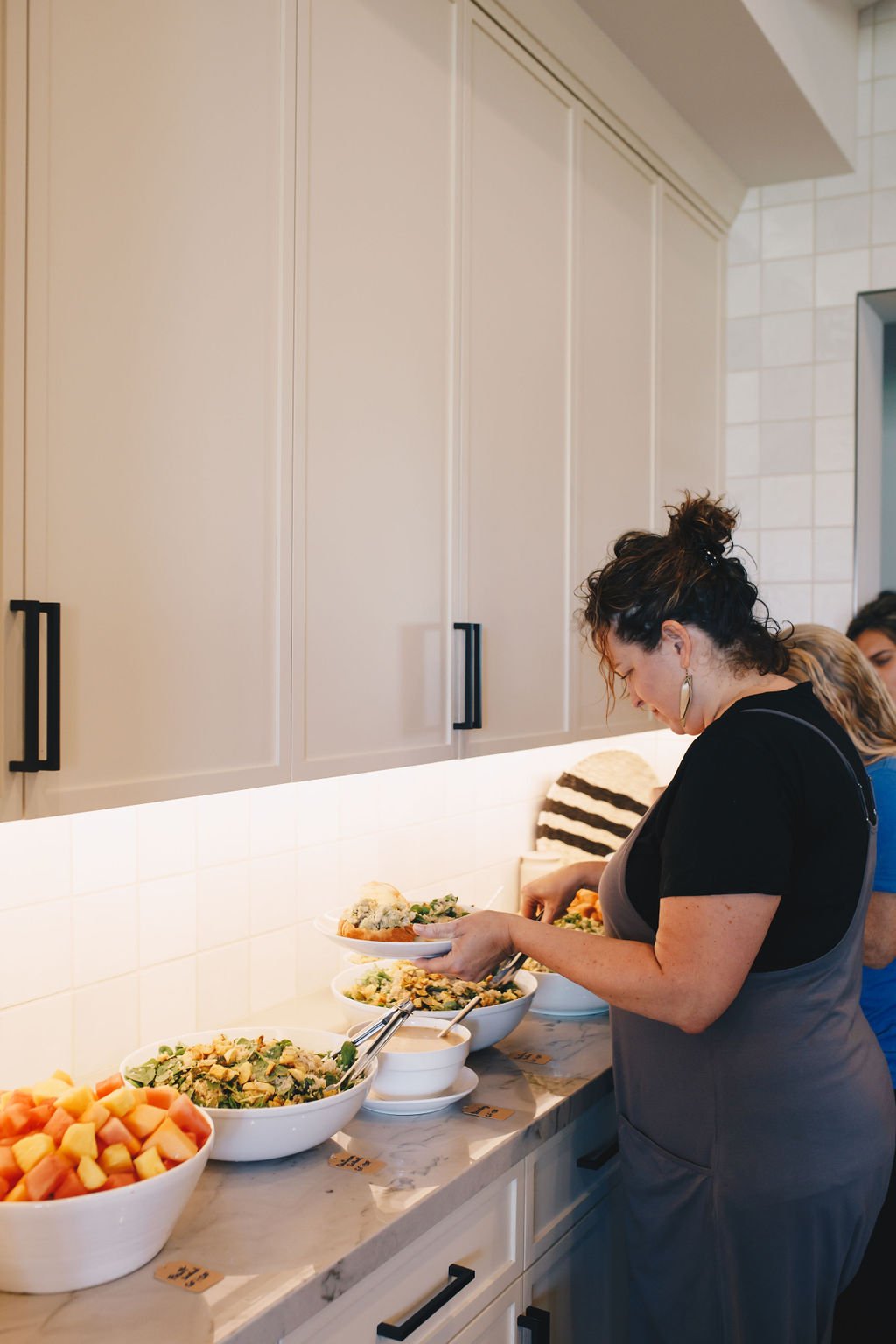 Woman serving herself from a variety of bowls of salad and fruit on a kitchen counter.