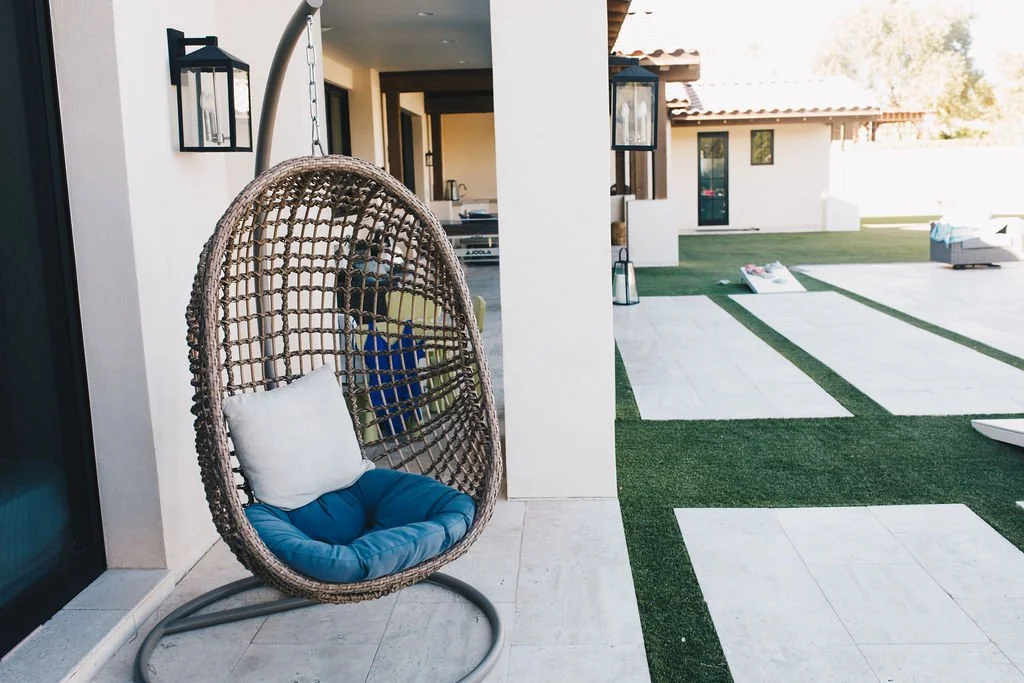 Outdoor patio with a hanging egg chair with blue cushion and white pillow, lanterns, and a view of a grassy yard with a white building in the background.